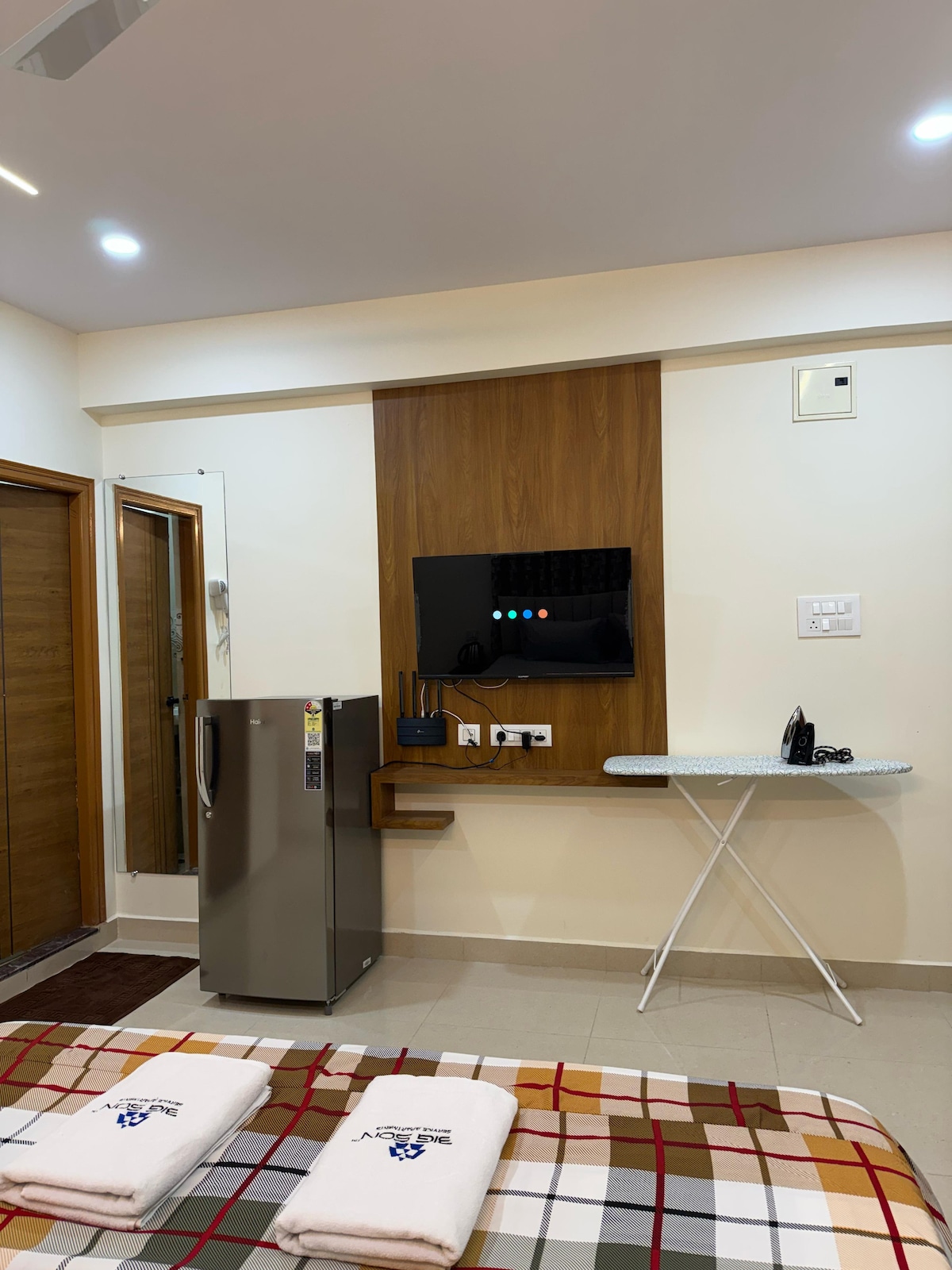 A well-furnished room featuring a mounted flat-screen television above a sleek wooden panel. A refrigerator stands nearby, complemented by a small table and ironing board. Minimalist decor creates a clutter-free environment, with light colors enhancing the spacious feel of the area.