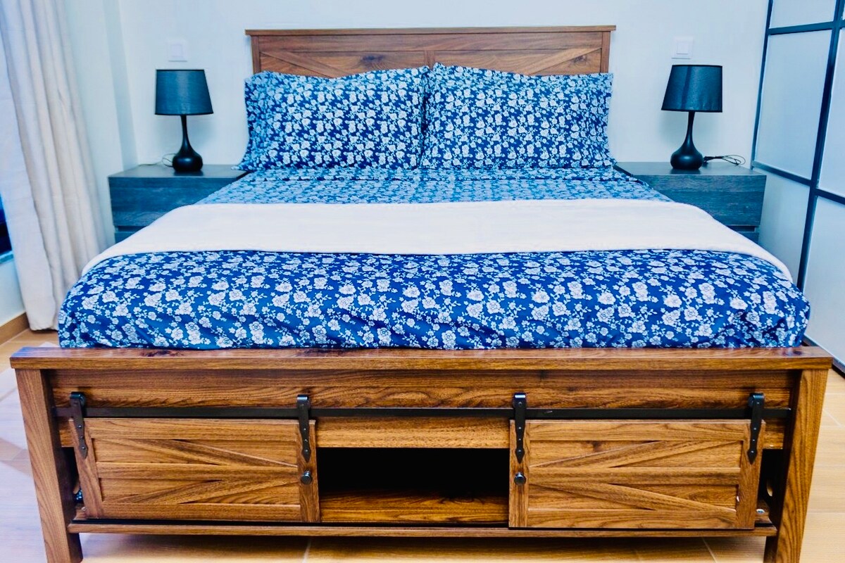 A spacious bed with a wooden frame is dressed in a floral-patterned blue duvet. Two bedside lamps with black shades provide soft lighting. The bed is positioned against a light-colored wall, with a wooden storage unit visible underneath.