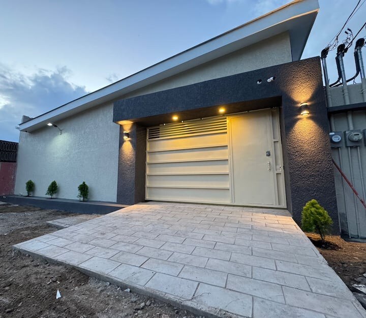 The exterior of the modern accommodation presents a sleek facade with contrasting light and dark colors. A spacious driveway leads up to a prominent garage door, which is illuminated by outdoor lighting. Small plants are situated along the concrete pathway, enhancing the overall appeal.