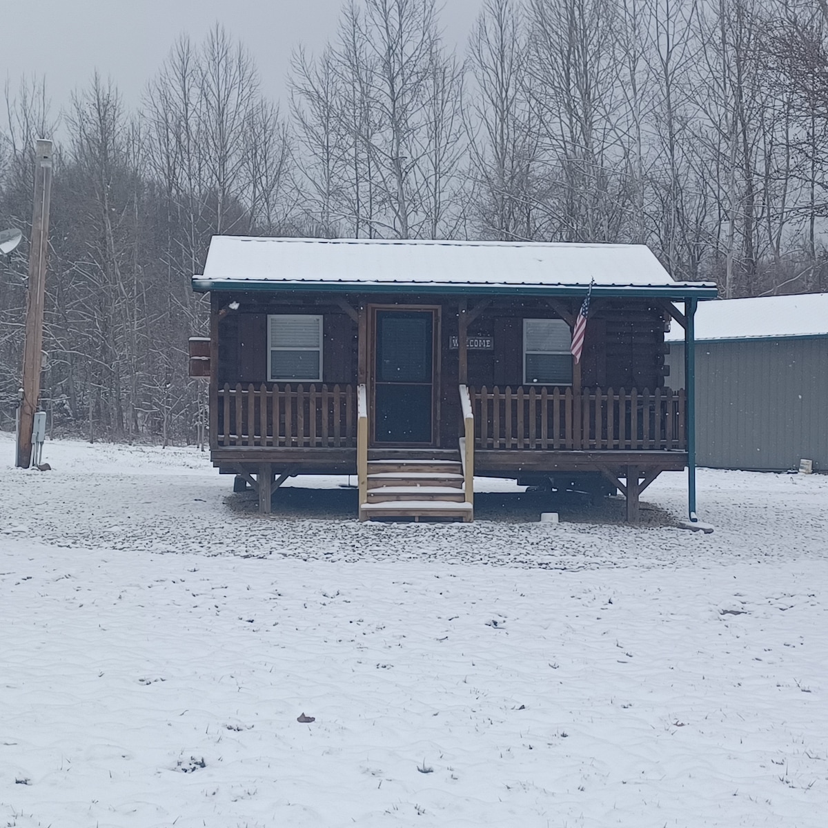 The rustic cabin features a front porch with a wooden railing, providing a welcoming entrance. Snow blankets the ground, and surrounding trees are visible. An American flag is displayed at the front, adding to the cabin's charm.