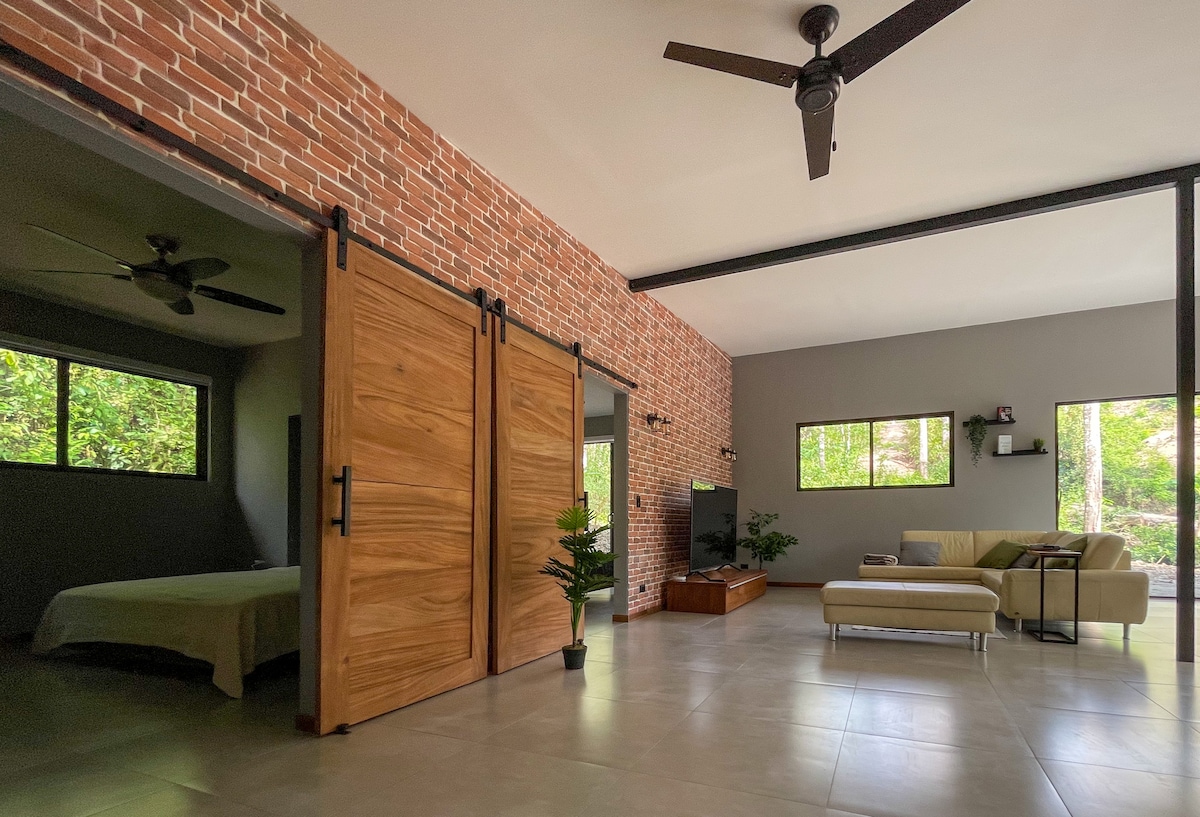 The living area features an open layout with large windows allowing natural light. A comfortable sofa is positioned near a wall-mounted TV. Adjacent, sliding wooden doors lead to a bedroom, while a small plant adds a touch of greenery.
