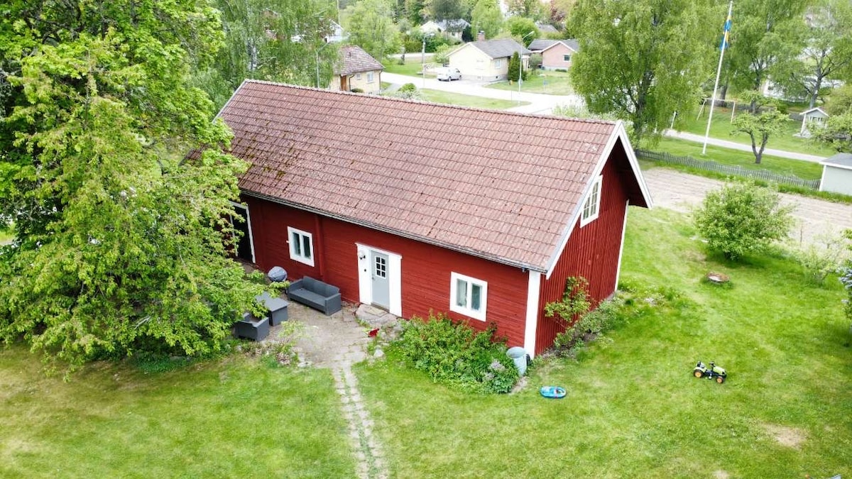 The charming red house is surrounded by a lush green lawn, featuring a stone pathway leading to the entrance. Outdoor seating is arranged on the patio, nestled under the shade of a tree. Nearby, children’s toys are seen, indicating a family-friendly environment.