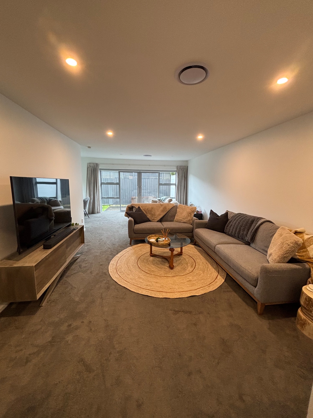 A modern living area is presented with a gray sofa and a comfortable armchair positioned around a circular coffee table. A flat-screen television sits atop a low console. Natural light enters through large sliding doors, which lead to a fenced outdoor area. A neutral rug enhances the space.