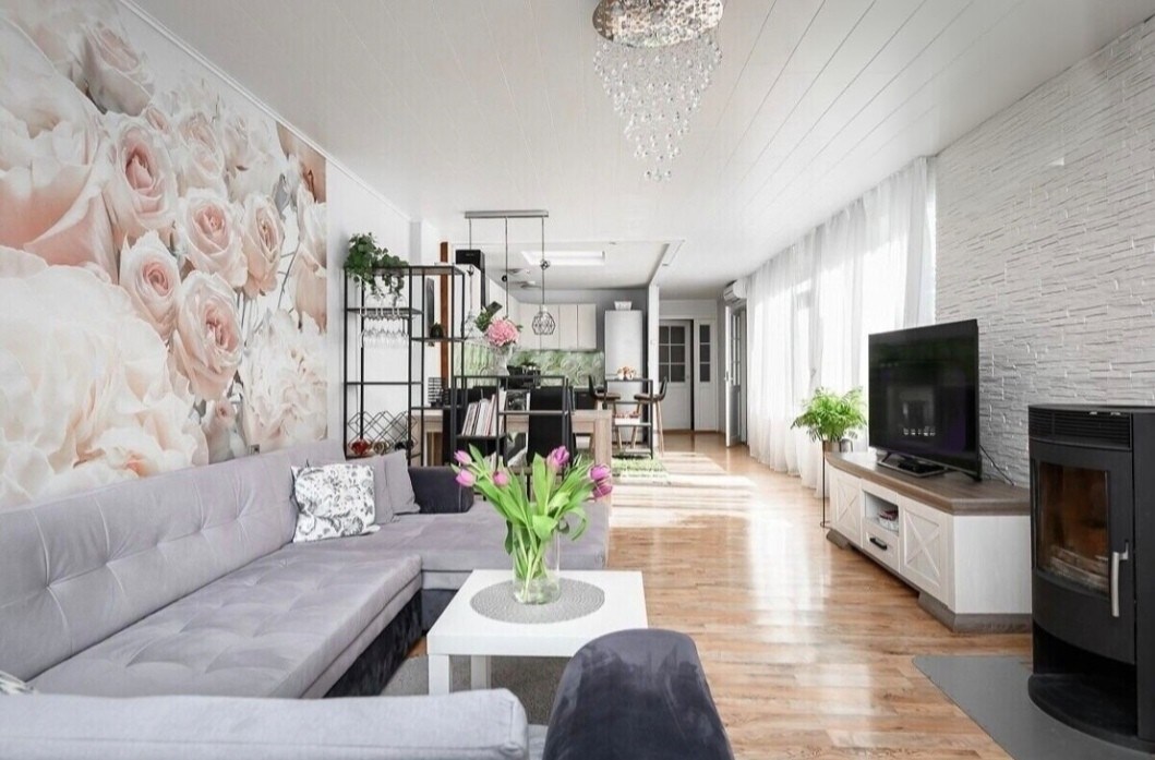 A spacious living area features a large sectional sofa in a soft gray hue, complemented by a coffee table. A vibrant floral wall mural and a modern chandelier add character to the room, while a fireplace and an entertainment unit are seen in the background.