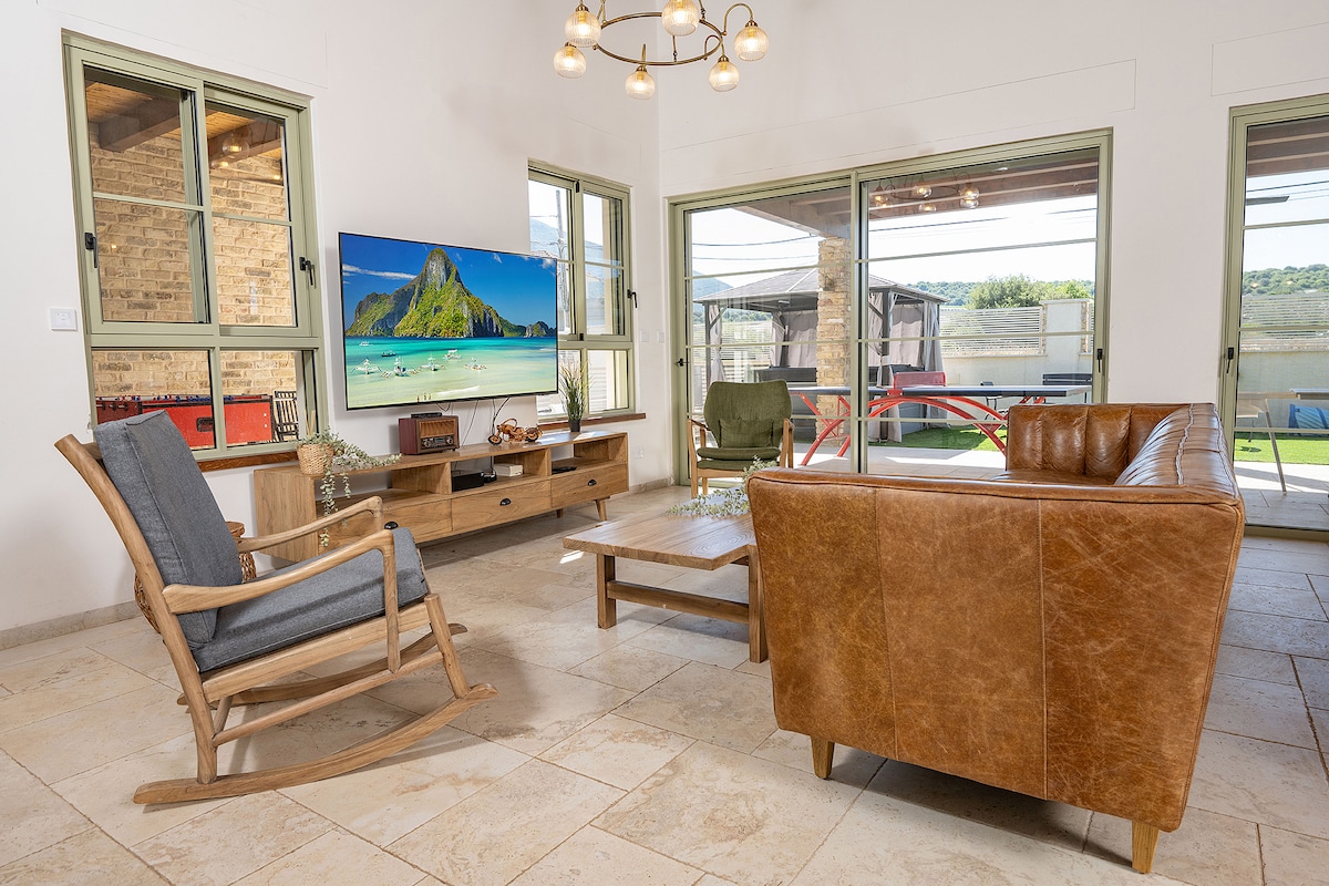 A living area is presented with large windows allowing natural light to fill the space. Comfortable seating includes a leather couch and a wooden rocking chair. A wooden coffee table is centered in the room, while a television mounted on the wall displays an image of a tropical landscape.