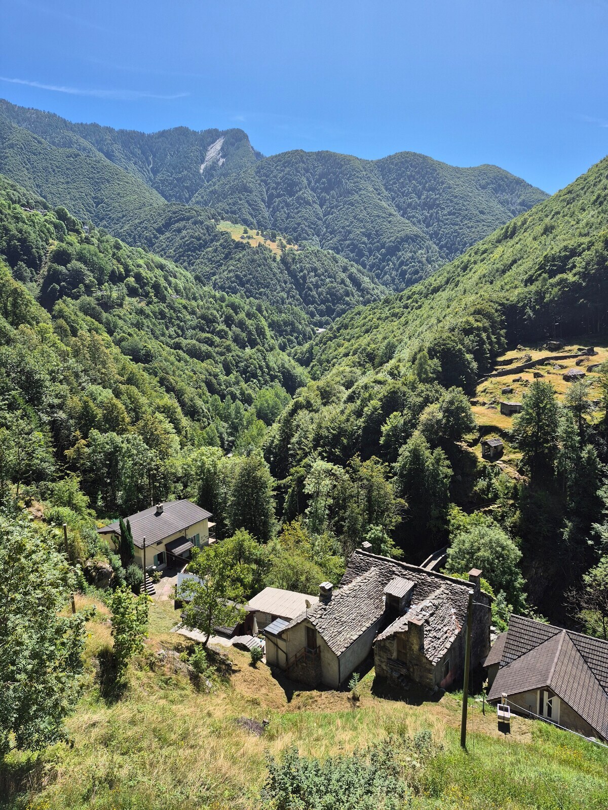 A serene landscape features lush green mountains and valleys under a clear blue sky. Traditional stone cottages are nestled amongst the trees, with open grassy areas visible. The scene embodies a peaceful retreat, ideal for nature enthusiasts seeking tranquility.