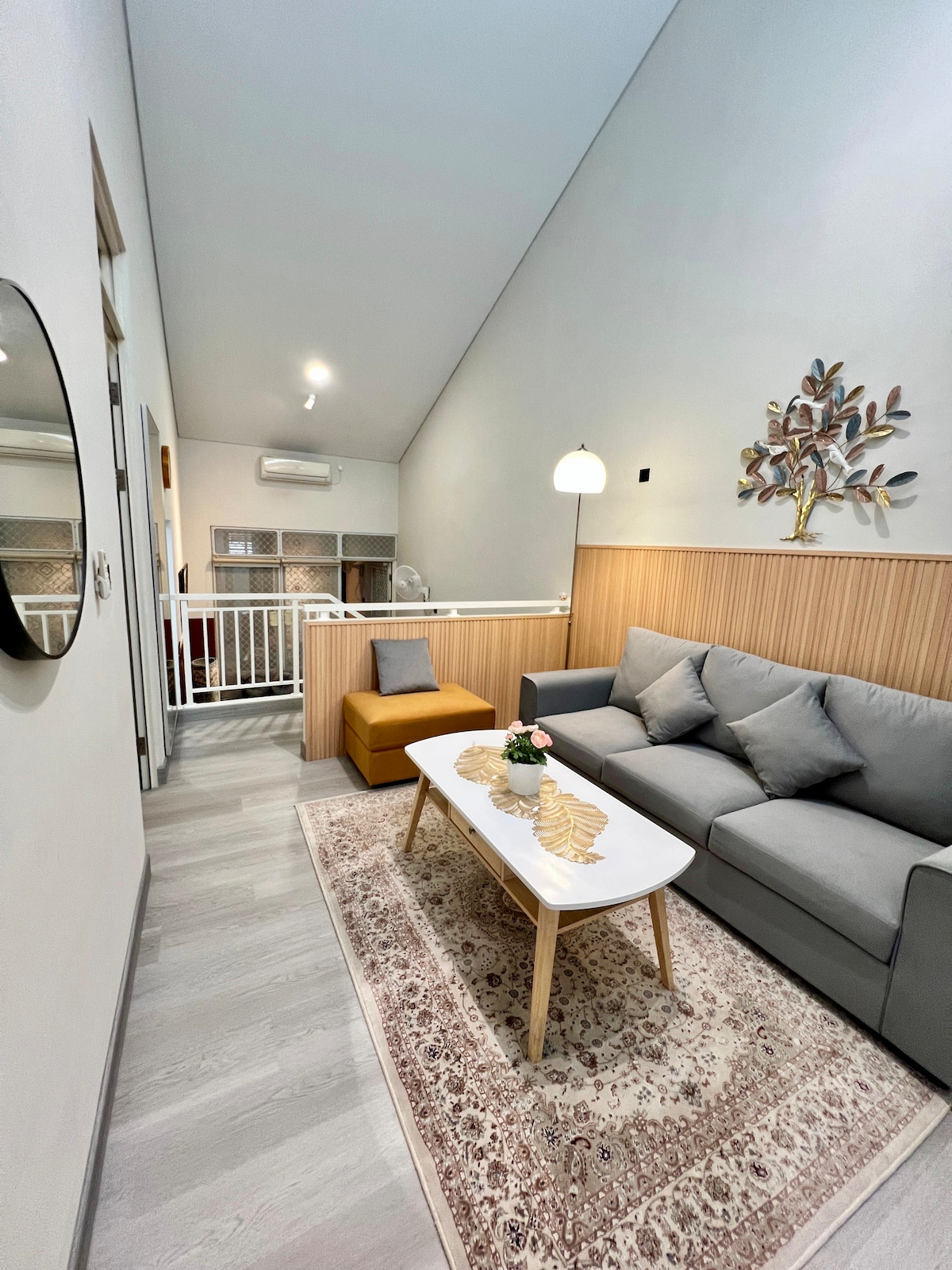 A well-lit living area showcases a gray sofa and an accent chair in a warm tone. A minimalist coffee table is adorned with a floral centerpiece. Light walls create an airy feel, complemented by a decorative wall art piece and a warm-colored area rug.