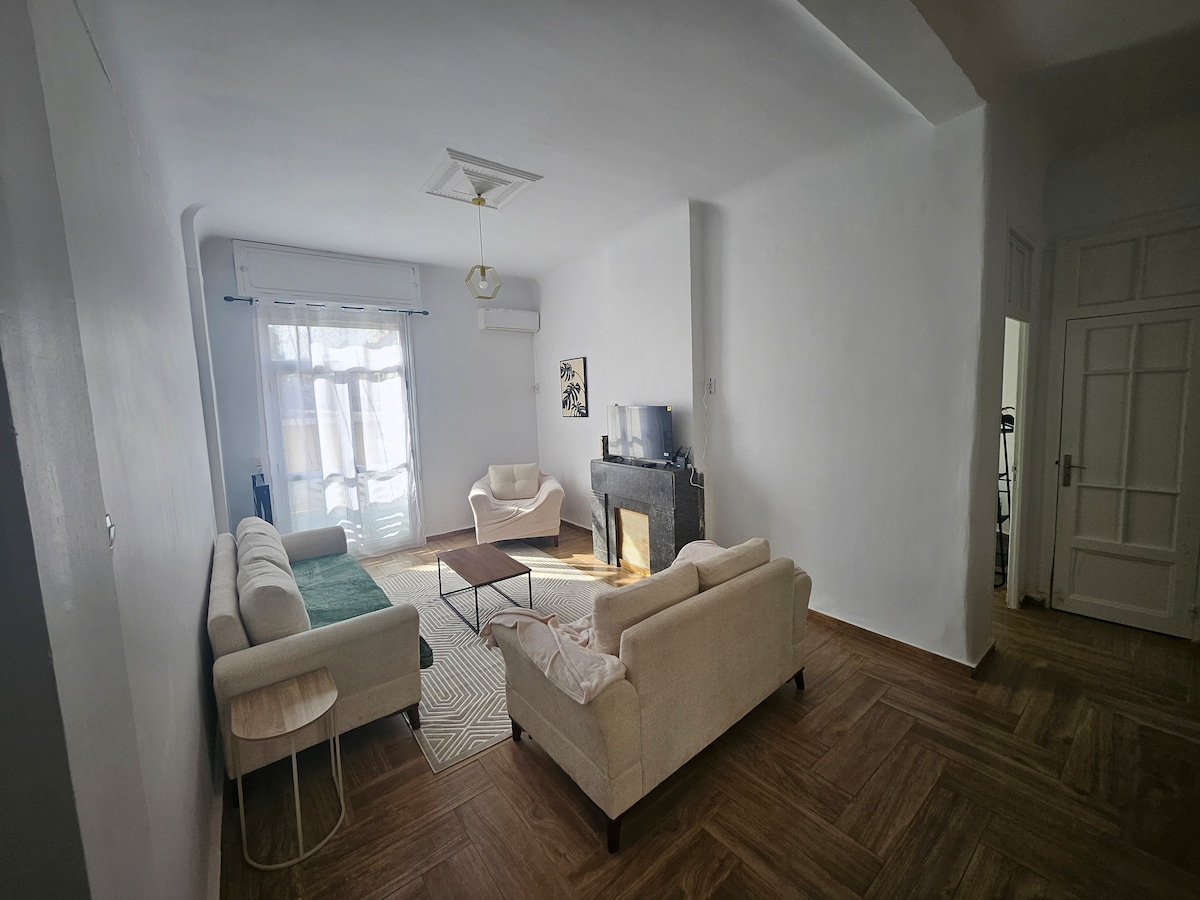 A spacious living area is furnished with two light-colored sofas and a small round side table. A flat-screen TV is positioned on a dark fireplace. Soft natural light enters through a large window, and a textured rug adds warmth to the wooden floor.