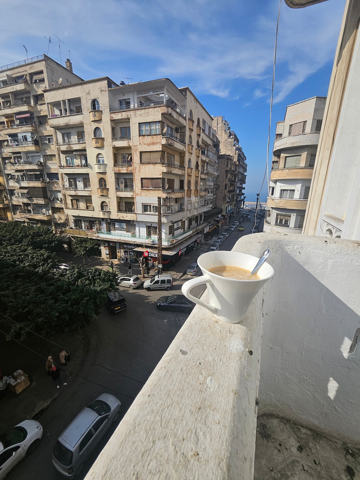 A view from an apartment balcony showcases an urban street scene with mid-rise buildings lining the road. A white mug sits on the balcony ledge, filled with coffee, while the blue sky hints at nearby sea visibility in the distance.