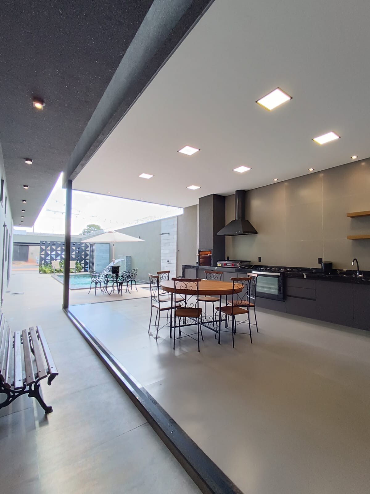 An open-concept kitchen and dining area is visible, featuring a large table surrounded by metal chairs. The sleek kitchen includes modern appliances and cabinetry. Large windows provide natural light and a view of the outdoor space, which showcases a dining set under an umbrella.
