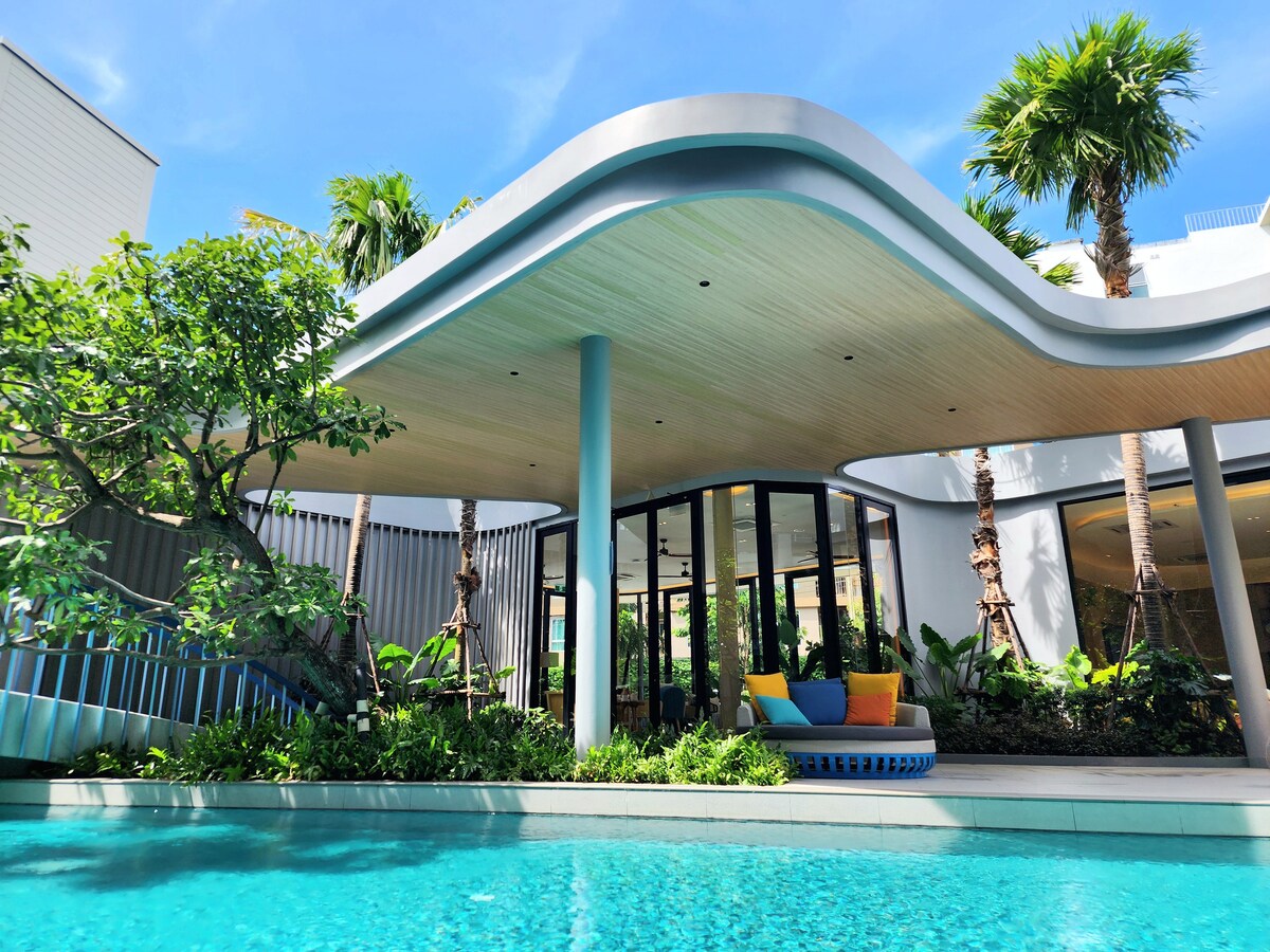 A modern architectural design is highlighted with a curved roof, surrounded by lush greenery. Large glass doors provide views of the bright interior, while a vibrant seating area is positioned by the pool, offering a serene outdoor space.