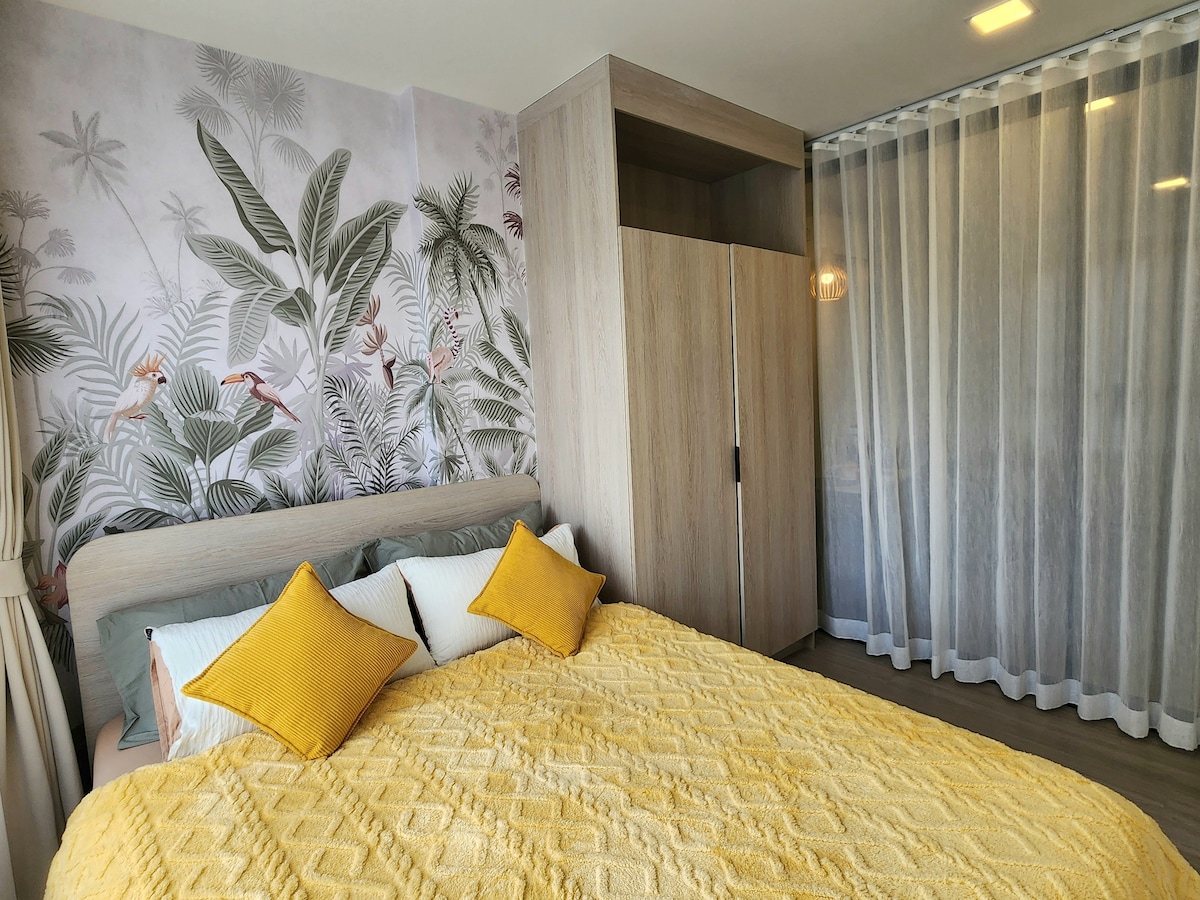 A soothing bedroom space features a spacious bed adorned with a textured yellow blanket and coordinating decorative pillows. Behind the bed, a botanical wallpaper creates a calming backdrop, and a light wooden wardrobe stands nearby, accompanied by sheer curtains that allow soft natural light to filter in.