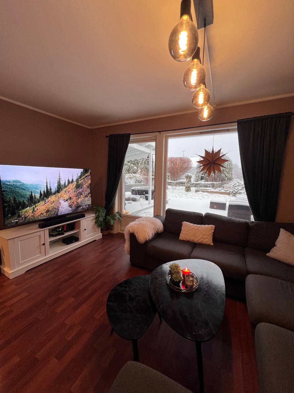 A cozy living area features a large dark sectional sofa and two stylish black coffee tables. A flat-screen TV rests on a white media unit against a brown wall. Natural light enters through large windows, partly obscured by dark curtains, while a decorative star-shaped light fixture hangs above.