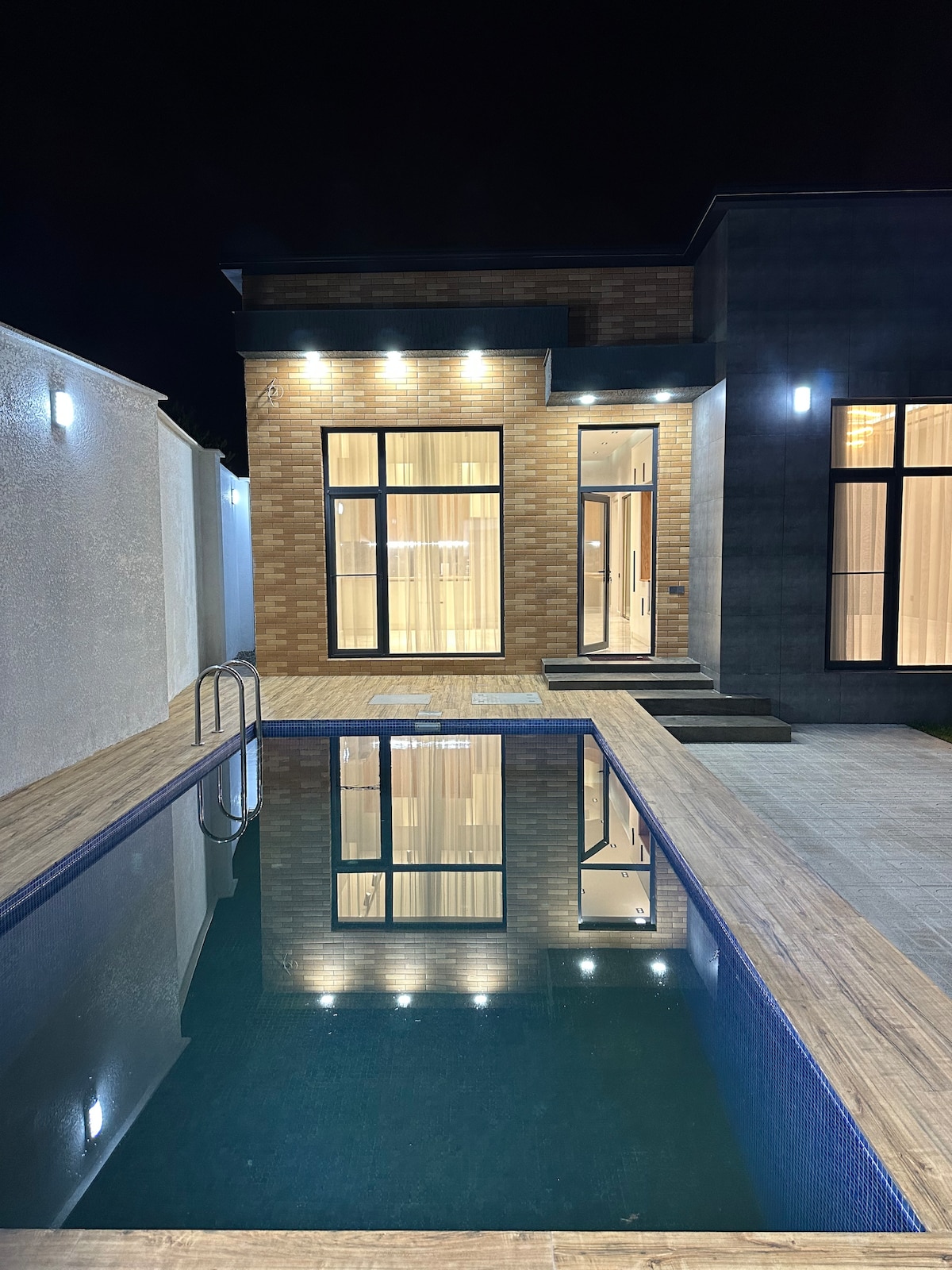 A modern exterior view of the home is presented at night, showcasing a sleek design with large, illuminated windows. A private pool reflects the building, surrounded by wooden decking, which enhances the serene ambiance of the outdoor space.