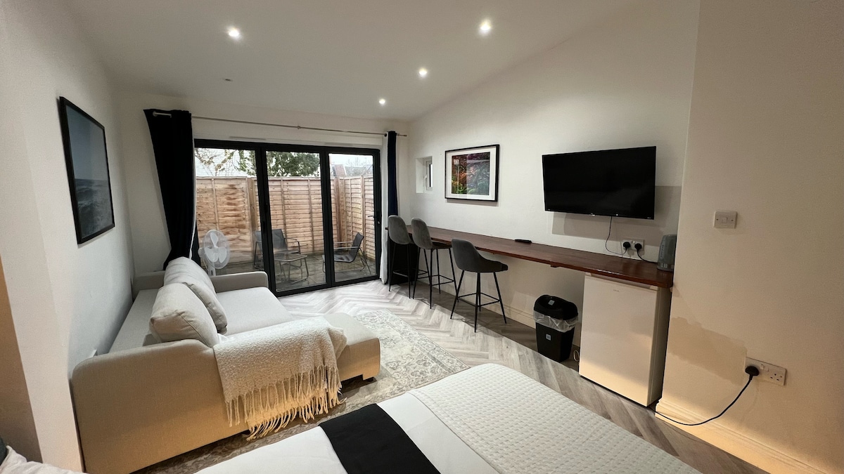 A spacious studio is shown, featuring a comfortable sofa and a television mounted on the wall. Large glass doors open to a private garden area. A dining space with high chairs and a sleek desk complements the modern design, illuminated by overhead lighting.