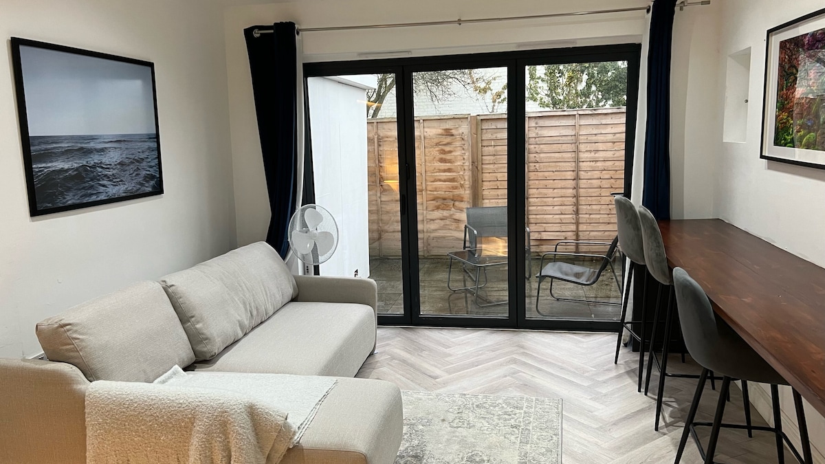 A light-filled living area is presented, featuring a comfortable sofa facing large glass doors that open to a private garden. A wooden table with several chairs can be seen in the background. The walls are adorned with framed artwork, adding a touch of character.