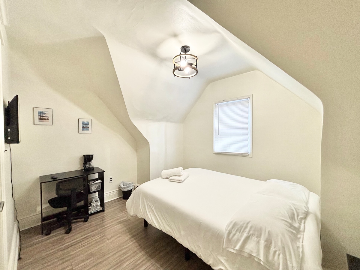 A room with an angled ceiling, featuring a spacious bed covered with a white blanket. A small desk with a chair and a coffee maker are positioned against one wall. A wastebasket is visible, and natural light enters through the window adorned with blinds.