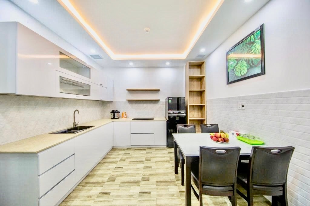 A functional kitchen area is highlighted by sleek, white cabinetry and modern appliances. A dining table with black chairs is set up for meals. Warm lighting is provided by recessed ceiling lights, complementing the light-coloured walls and wooden flooring.