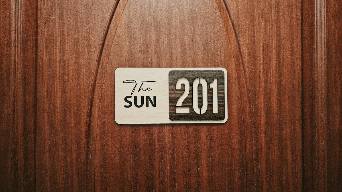 An inviting wooden door displays a stylish room number sign. The sign reads 'The Sun' above the number '201' in a contrasting color, set against the warm, rich texture of the door.