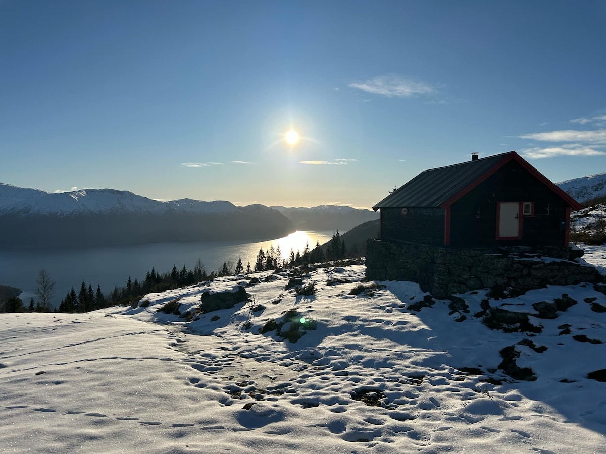 A rustic cabin is situated on a snow-covered hillside, offering a panoramic view of the Sognefjord. Sunlight reflects off the water's surface, enhancing the tranquil scene. The surrounding landscape features gentle slopes and distant mountains, creating a serene atmosphere.