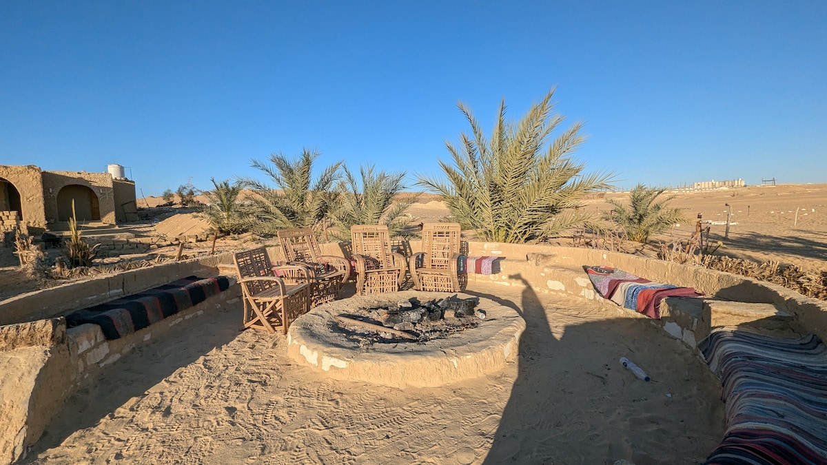 A circular seating arrangement surrounds a fire pit, with wooden chairs placed on a sandy surface. Colorful woven blankets add warmth to the area. Palm trees are visible in the background against a clear blue sky, creating a relaxed outdoor atmosphere.