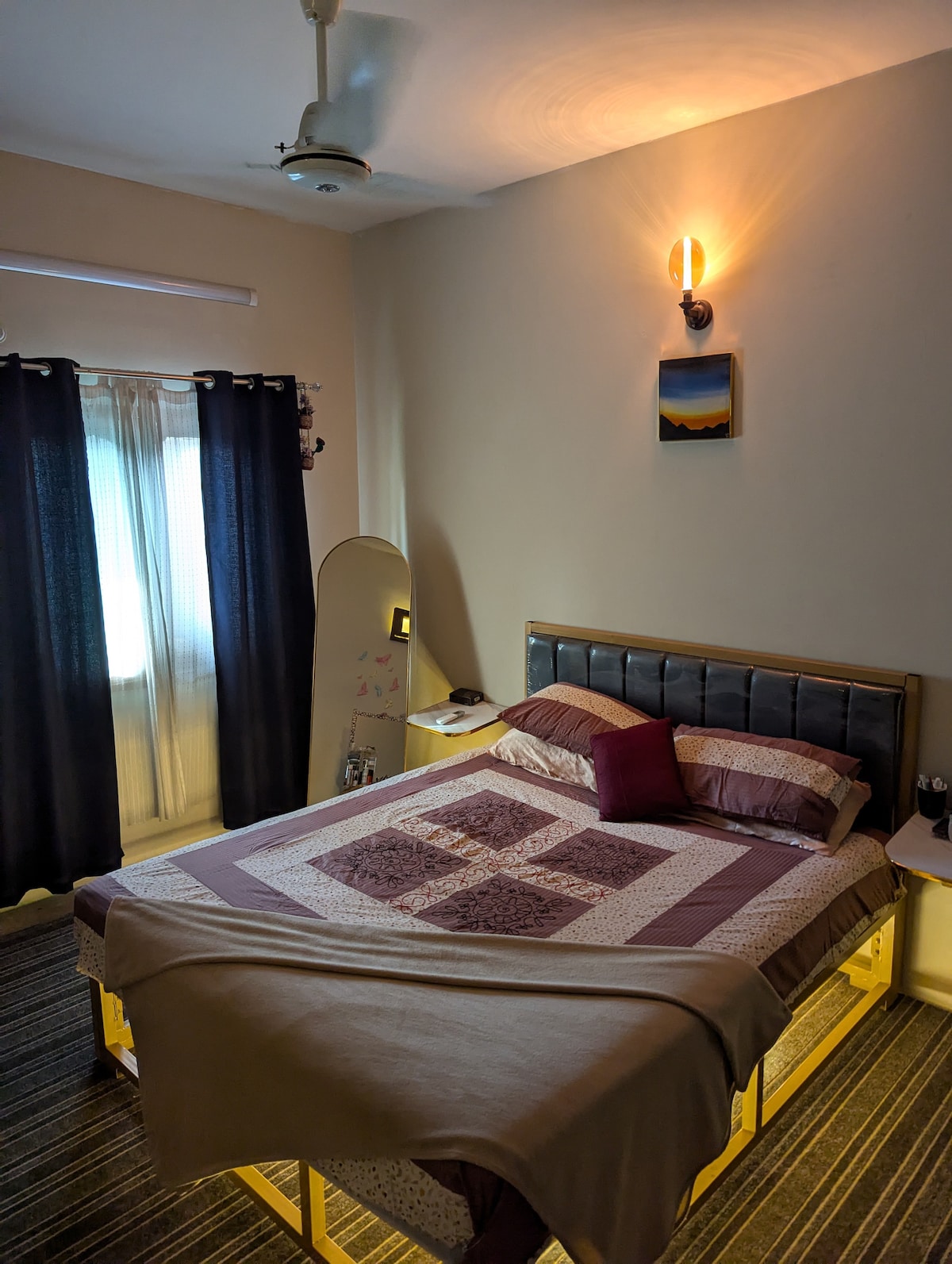 A tranquil sleeping area features a spacious bed with a patchwork quilt and coordinating throw pillows. Soft lighting from a wall fixture illuminates the room, while sheer curtains frame a window allowing natural light. The flooring consists of dark, striped carpeting, adding warmth to the space.