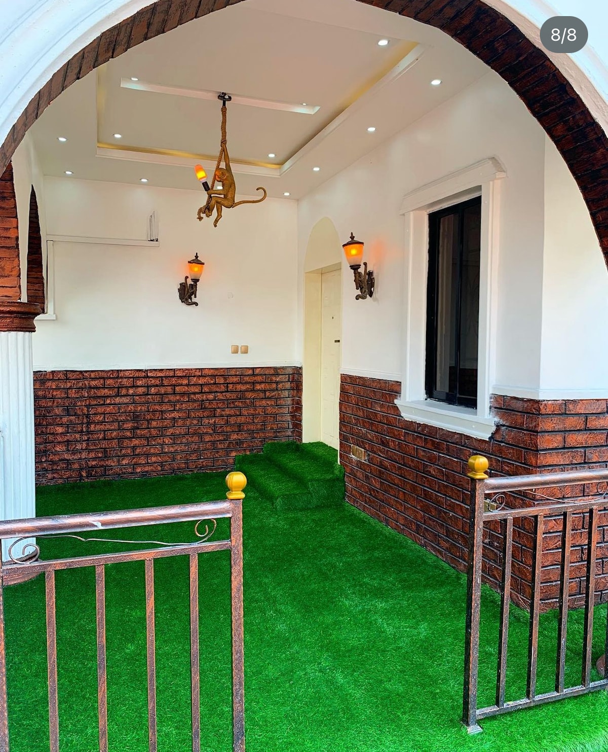A charming entrance area features an elegant archway with decorative lighting. Lush green artificial grass covers the floor, complemented by brick walls. A staircase leads to an interior door, while ornamental wall sconces provide soft illumination, enhancing the welcoming ambiance.