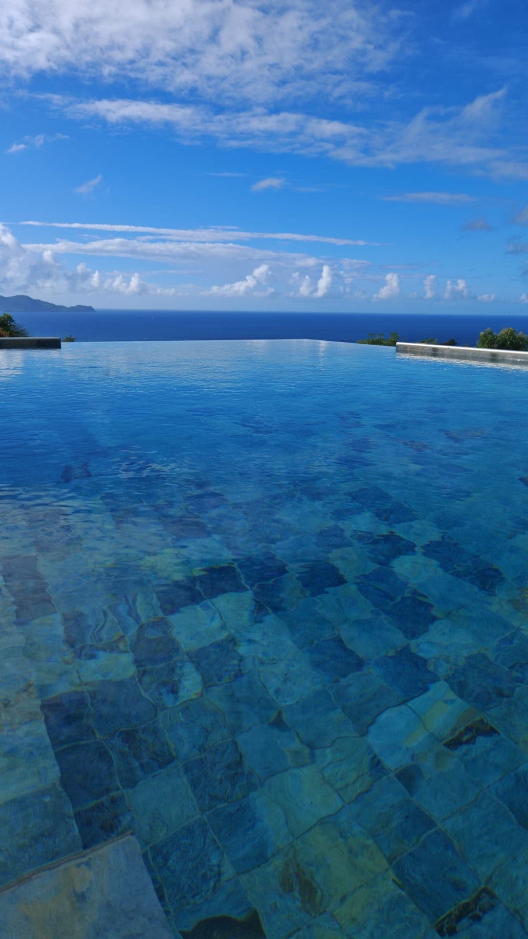 An infinity pool stretches seamlessly towards the horizon, offering unobstructed views of the Caribbean Sea. The water reflects a vibrant blue sky adorned with white clouds, creating a peaceful atmosphere. Lush greenery borders the pool, enhancing the tranquil setting.