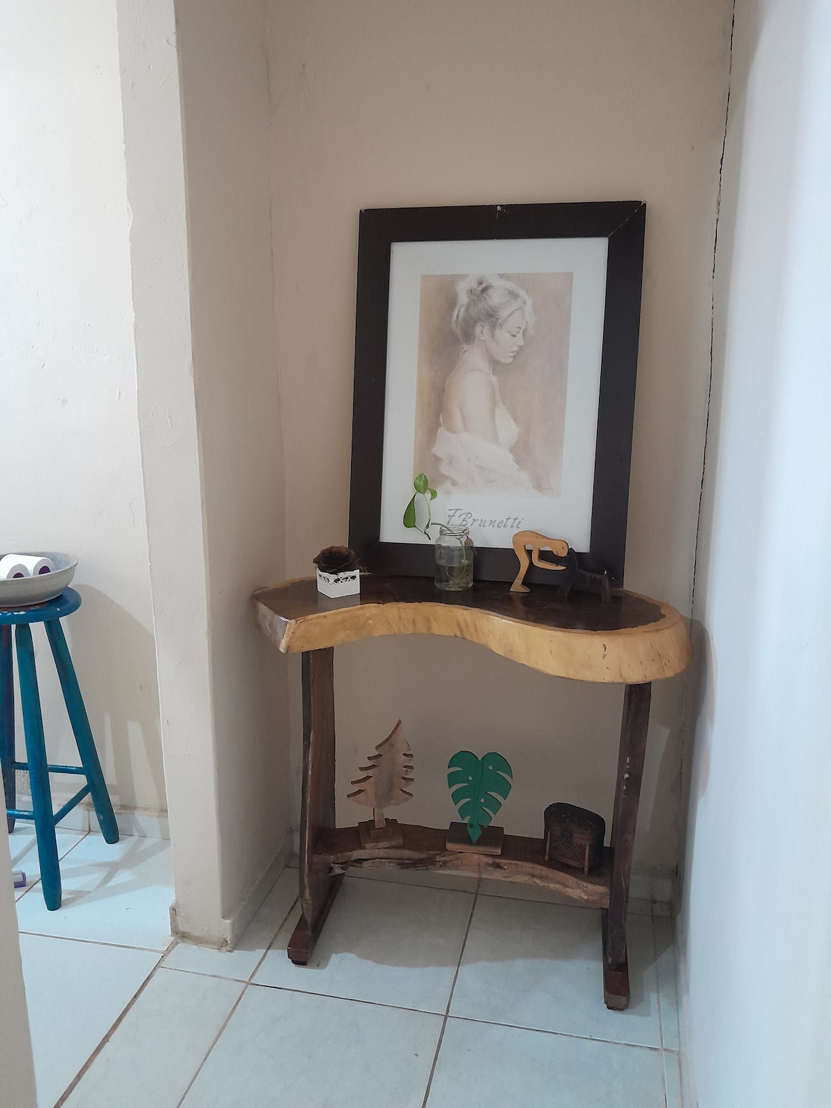 A corner table crafted from wood showcases natural design elements. A framed picture hangs above, featuring a portrait. A small glass container with foliage and a decorative item are placed on the table, contributing to the calm and stylish ambiance of the space.