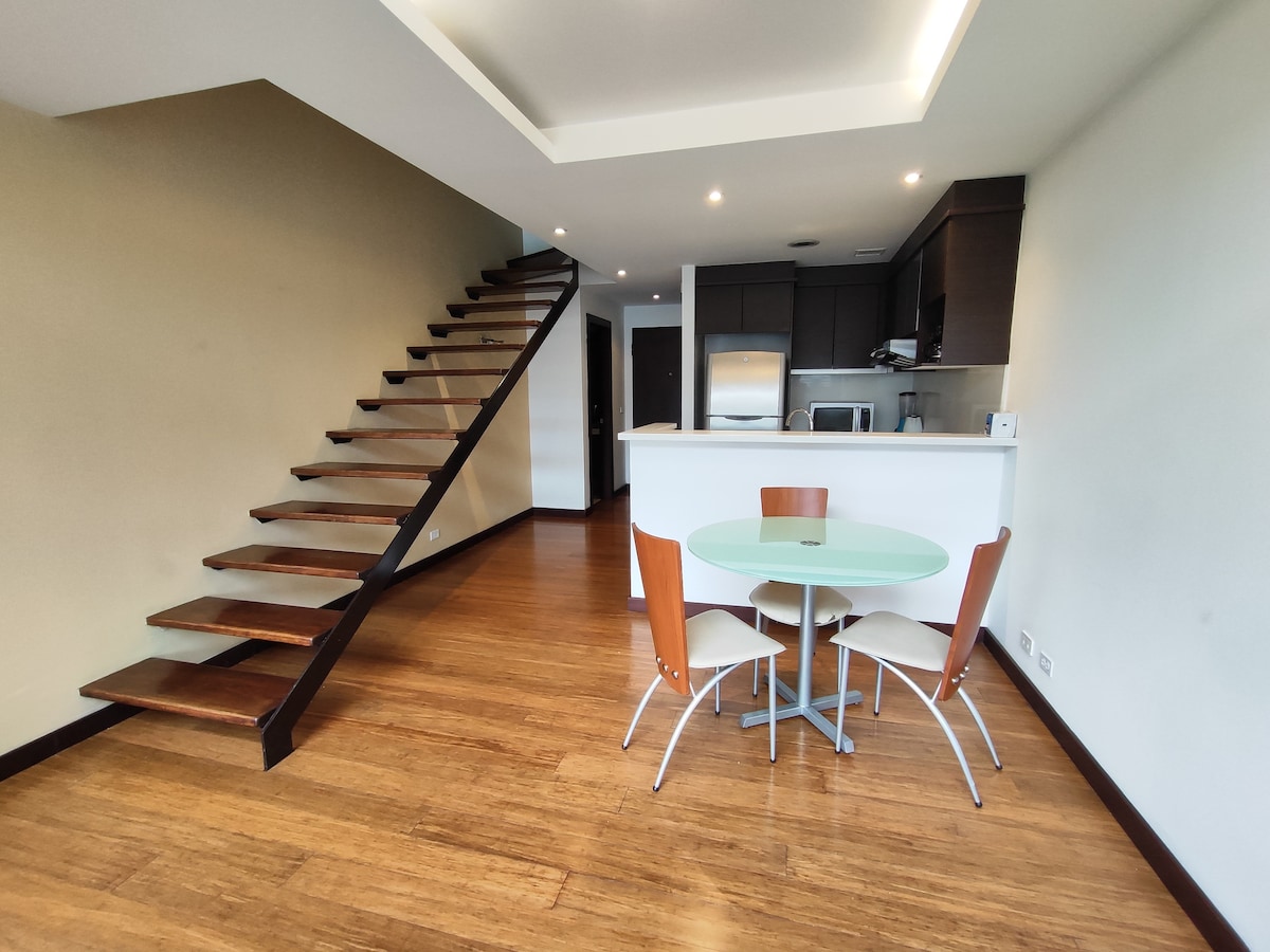 An open living area is presented with a modern spiral staircase leading to the upper level. A circular glass table is surrounded by four chairs, with a kitchen area visible in the background. Warm wood flooring complements the neutral-toned walls.