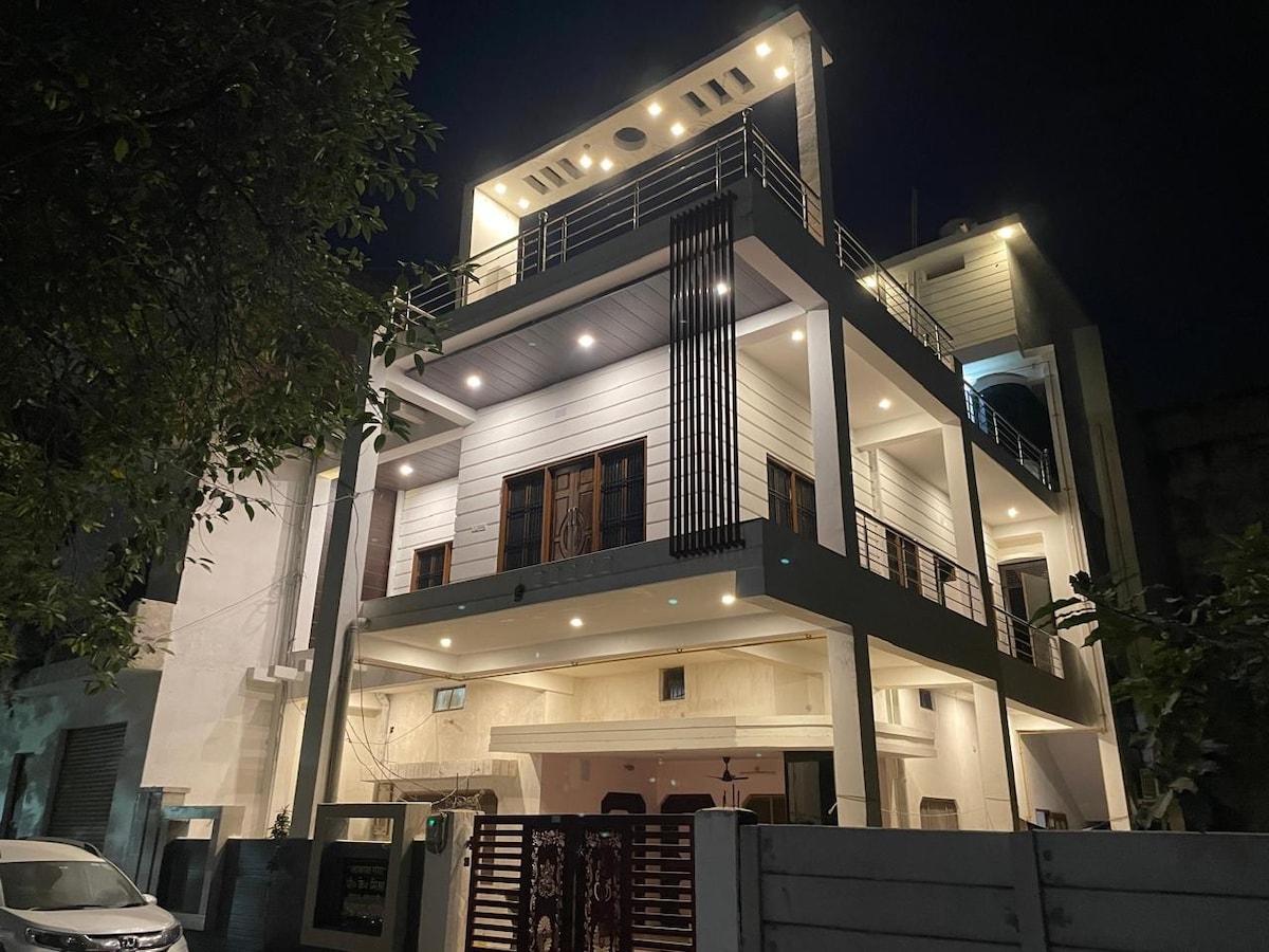 A three-story building is illuminated against the night sky, displaying a modern architectural design. The facade features a mix of white and wooden elements, along with spacious balconies. Soft lighting enhances the inviting appearance, while greenery is visible along the surrounding area.
