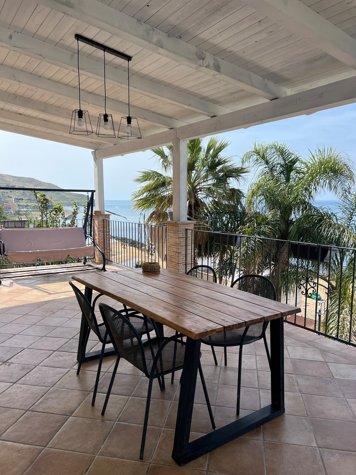 An outdoor dining area features a wooden table with black metal legs and six chairs, positioned under a covered terrace. Lush palm trees and a view of the sea are visible in the background, creating a serene environment for meals.