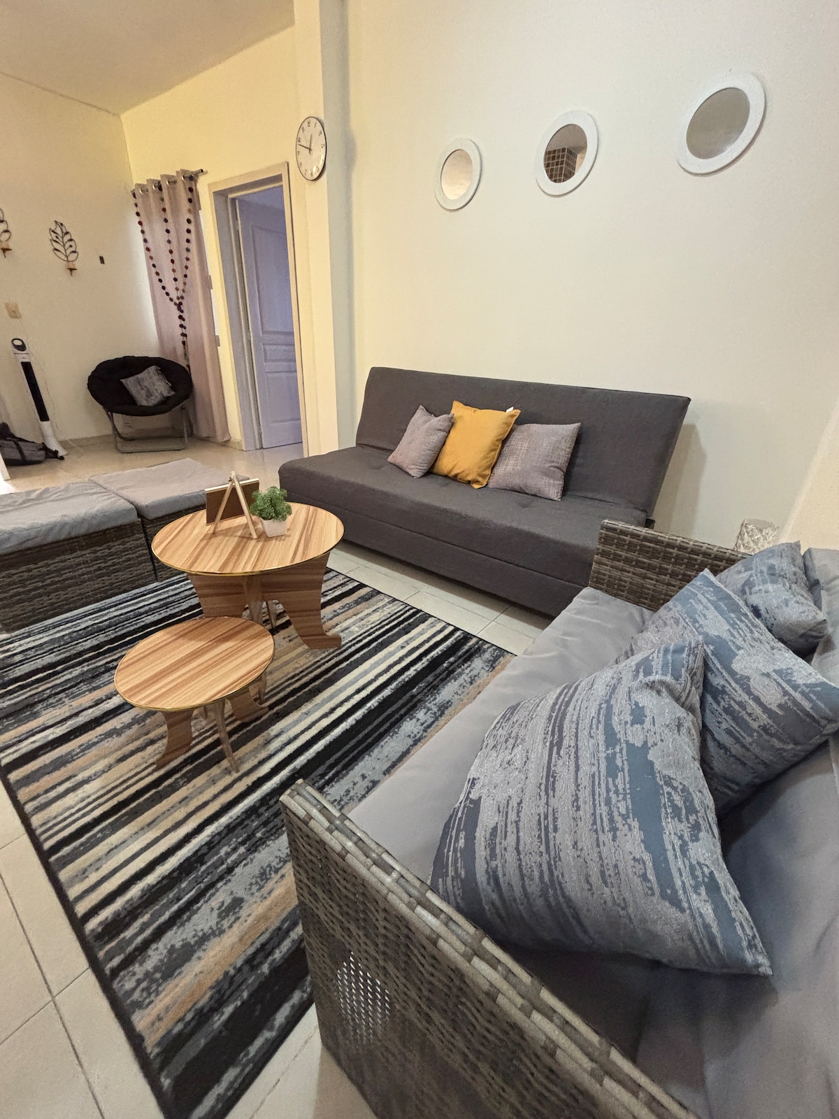 A modern living area is presented with a gray sofa adorned with orange and gray cushions. Two round wooden tables sit between comfortable wicker chairs. A striped rug anchors the space, while round decorative mirrors are mounted on the wall, enhancing the room's design.