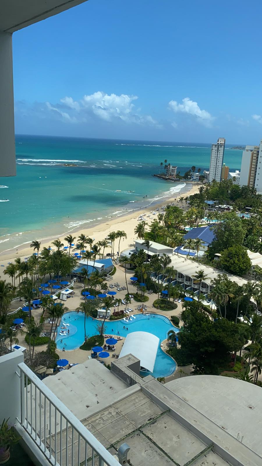 A panoramic view of a sandy beach is captured, highlighting the turquoise waters and palm trees. The scene includes a swimming pool area with seating under blue umbrellas. Beachfront buildings are visible in the background, contributing to the overall coastal atmosphere.