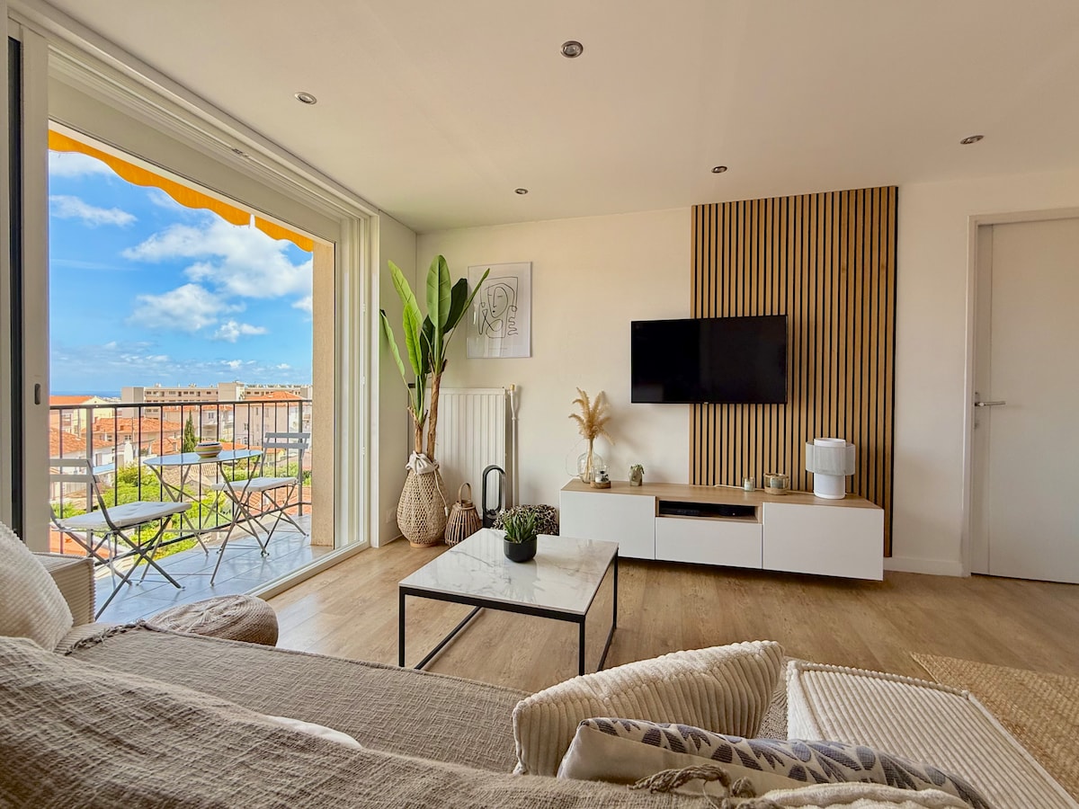 A light-filled living area features a modern entertainment unit with a television and a coffee table atop a wooden floor. A large window offers views of a balcony with outdoor seating while greenery adds freshness to the space.