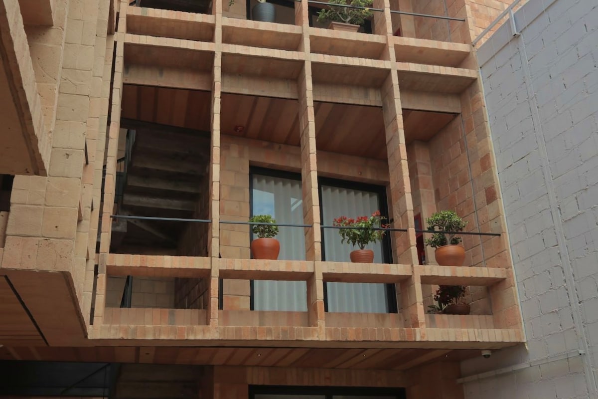 An exterior view of a modern building showcases a geometric facade featuring terracotta bricks and a grid-like balcony structure. Potted plants are seen on the balcony, adding greenery to the design. Large windows are framed by horizontal wooden beams.