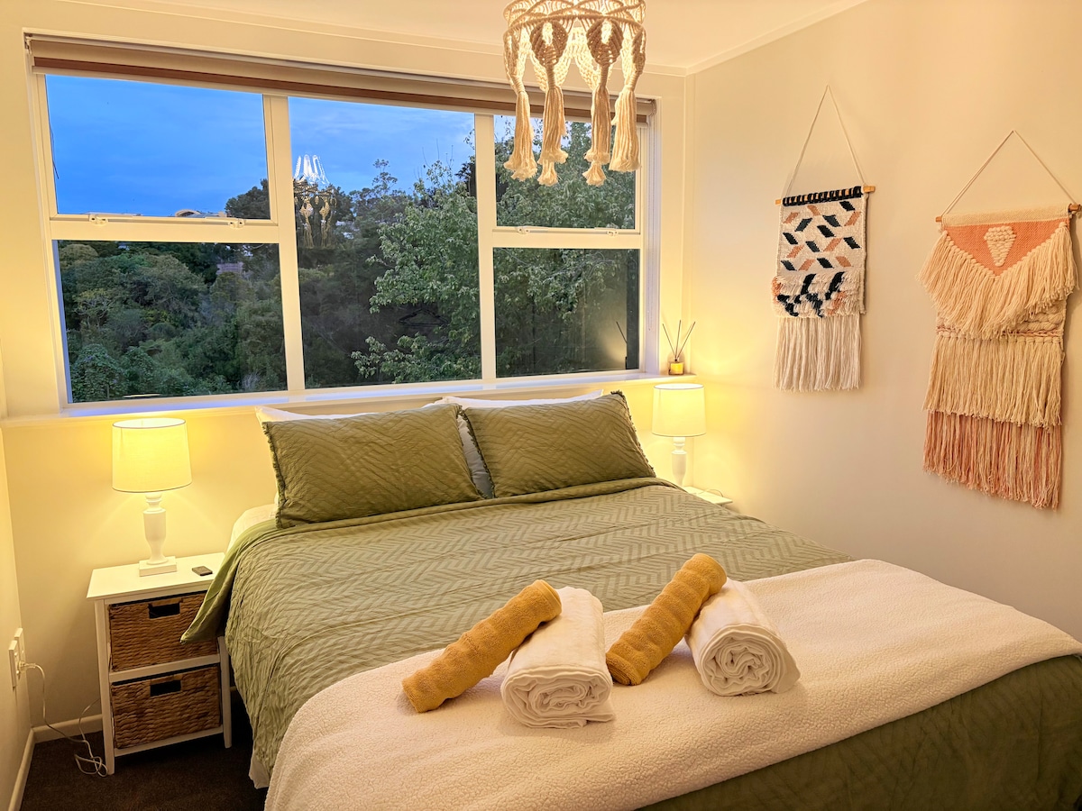 A cozy bedroom features a queen-size bed adorned with neutral-toned linens. Two bedside lamps offer soft lighting, and wall hangings add texture. Large windows showcase views of the surrounding greenery, allowing natural light to fill the space. Fresh towels are neatly arranged on the bed.