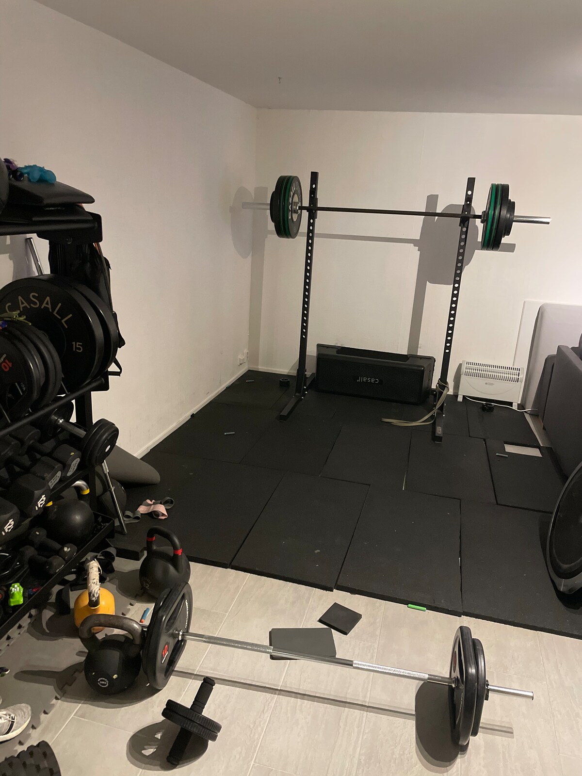 A dedicated workout space is equipped with free weights and a barbell. A squat rack stands against the wall, accompanied by rubber flooring for safety. The area is organized with various weight plates and exercise equipment, creating a functional environment for fitness activities.