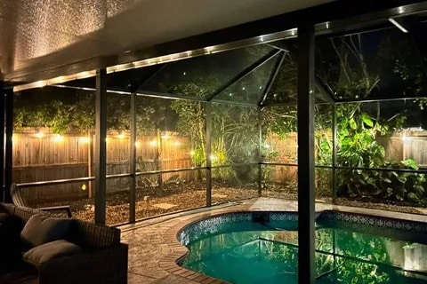 A fenced backyard is illuminated by soft outdoor lighting, creating a relaxed atmosphere. A heated pool is visible, surrounded by a paved patio area. Lush greenery borders the space, adding privacy and a natural element. Comfortable seating is arranged to enhance outdoor enjoyment.