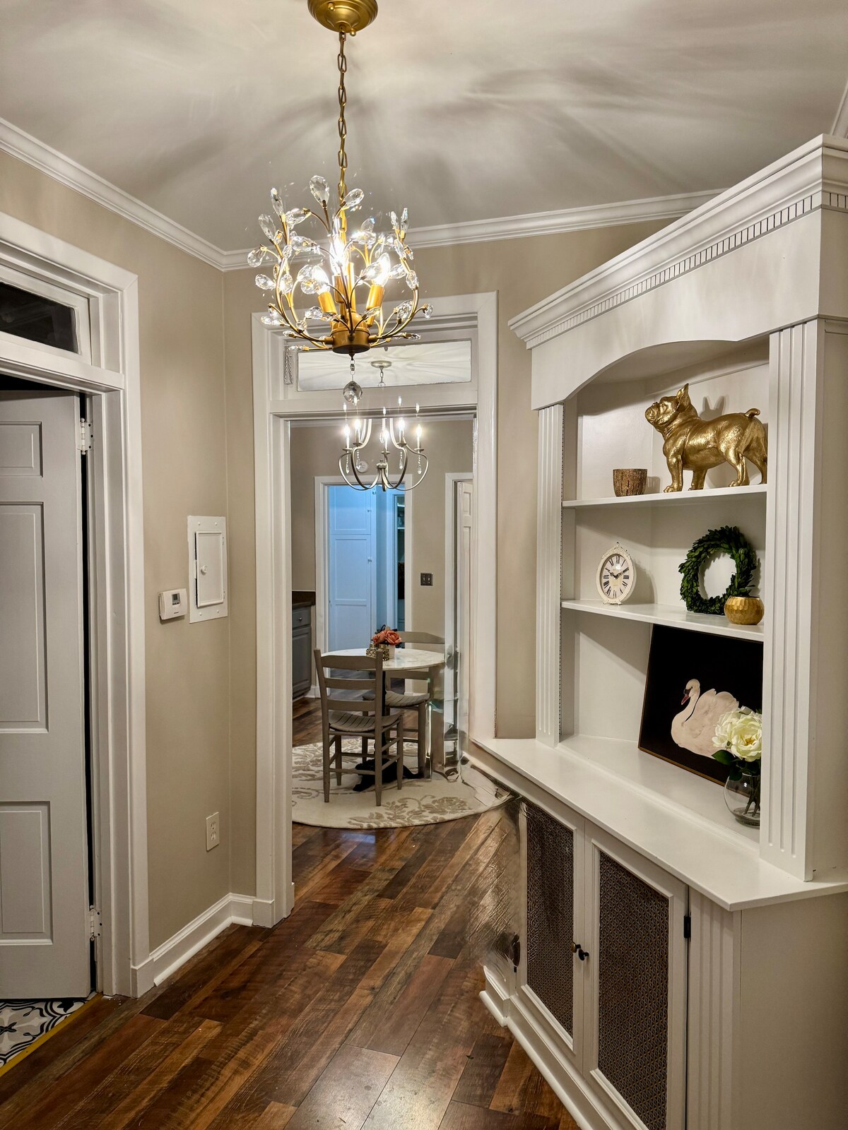 The hallway features light hardwood flooring and neutral walls, with a stylish chandelier casting soft light. A white shelving unit displays decorative items, and a round dining table is visible in the background, surrounded by chairs, inviting a cozy dining experience.