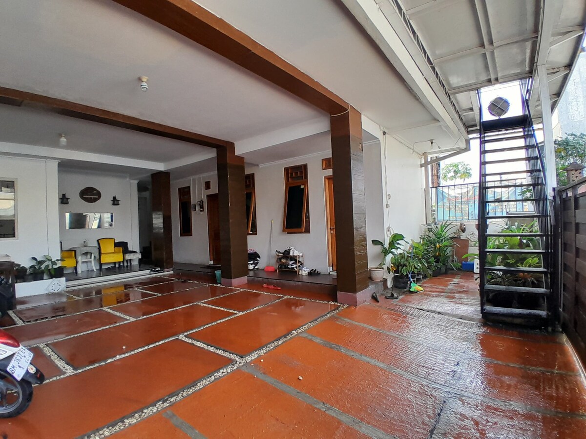 A covered entryway features a spacious, tiled floor with a combination of outdoor seating and potted plants. A staircase with a metal railing leads to an upper level. The area is illuminated by natural light, creating a welcoming atmosphere for guests.