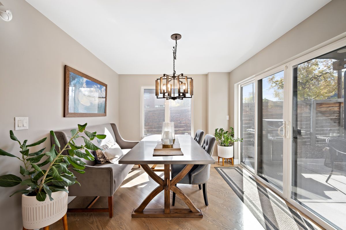 A dining area is presented with a long wooden table surrounded by dark chairs and light armchairs. Soft natural light enters from two glass doors, highlighting a potted plant and a simple decorative piece on the table. Neutral tones are used throughout the space.
