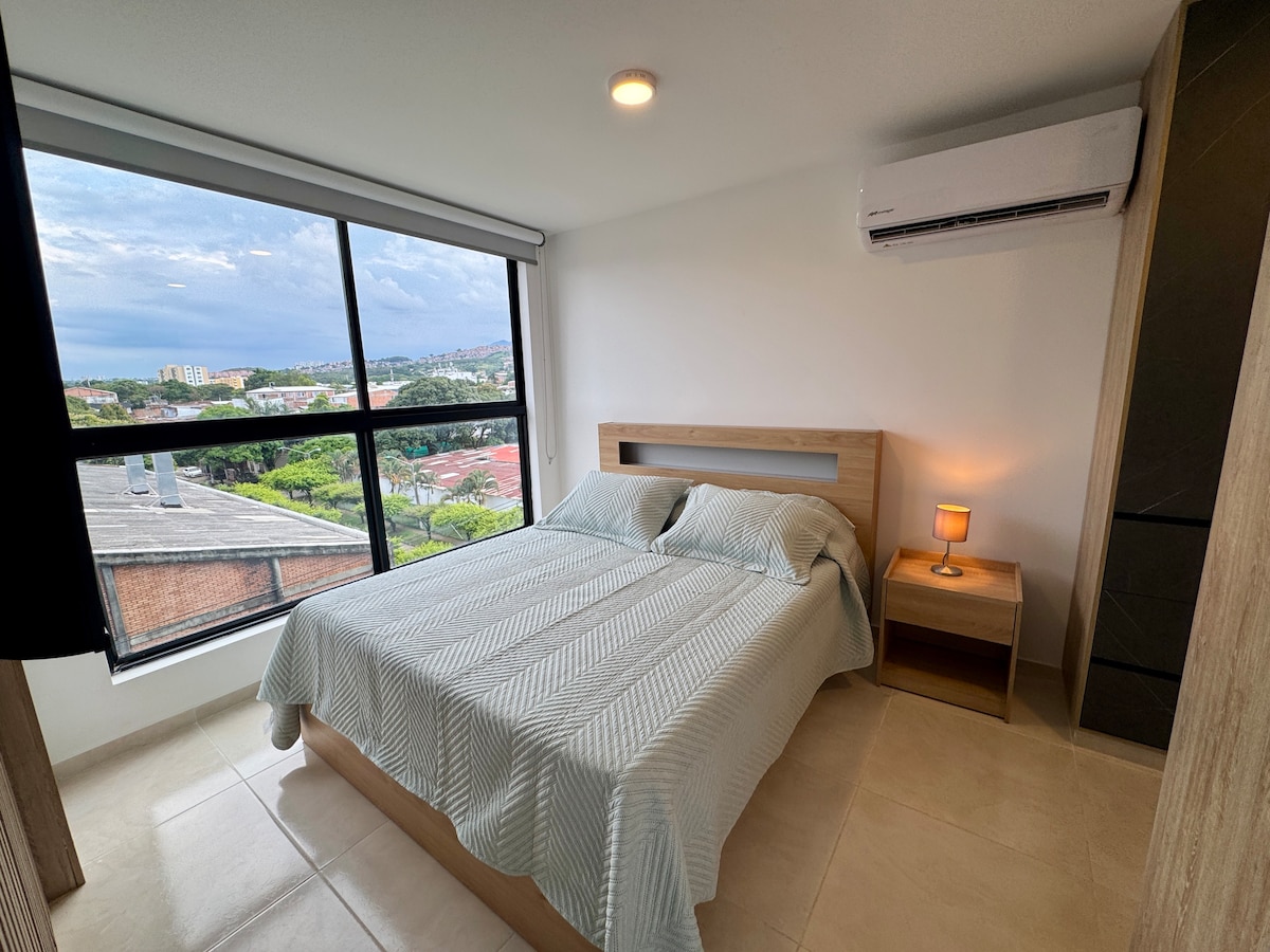 A well-lit bedroom features a spacious bed with a light-colored blanket, positioned against a large window that offers a city view. A bedside table with a lamp is next to the bed. An air conditioning unit is mounted on the wall above, enhancing comfort.
