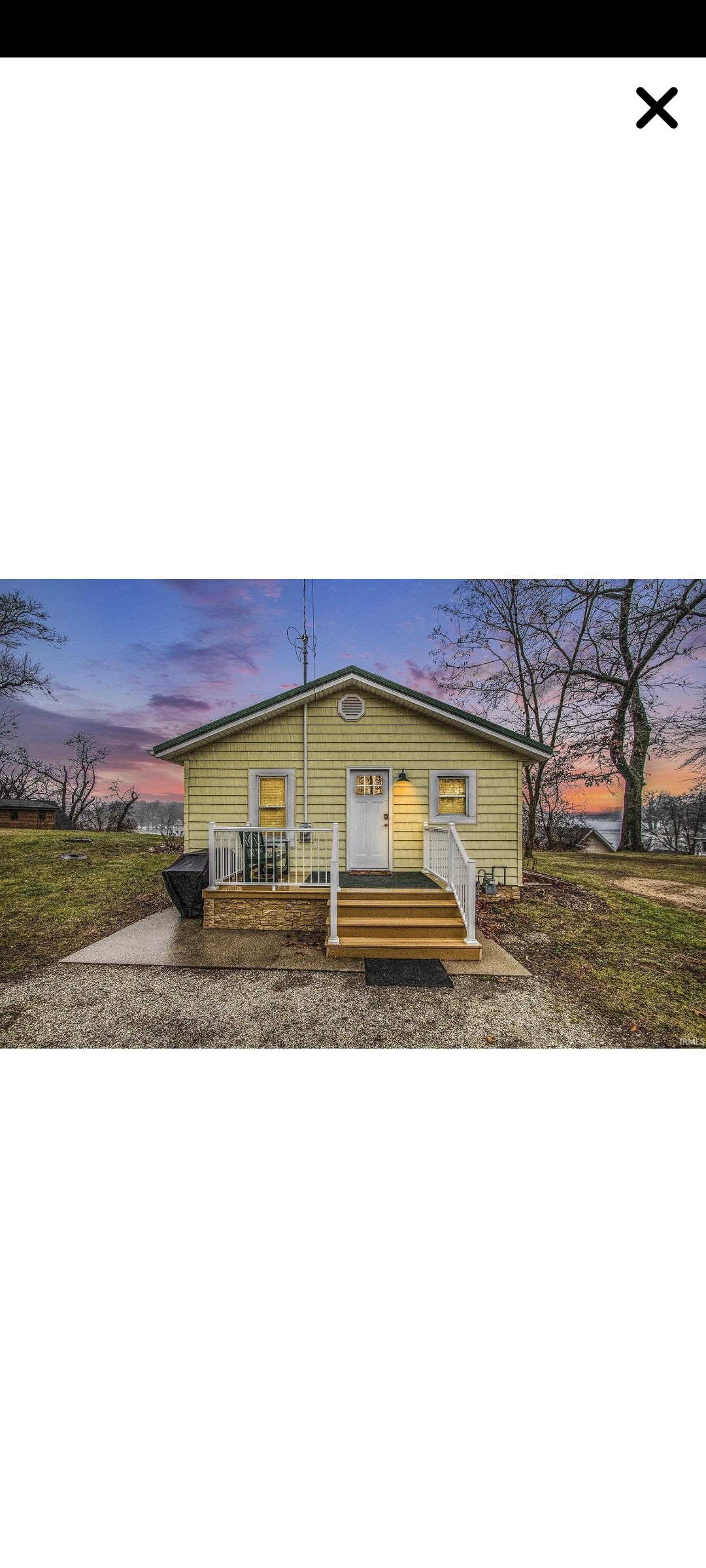A charming home is situated on a tranquil lot, featuring a welcoming front entrance with steps leading to a porch. Soft hues of dusk illuminate the exterior, highlighting the yellow siding and nearby trees, while a barbecue grill is positioned on the side.