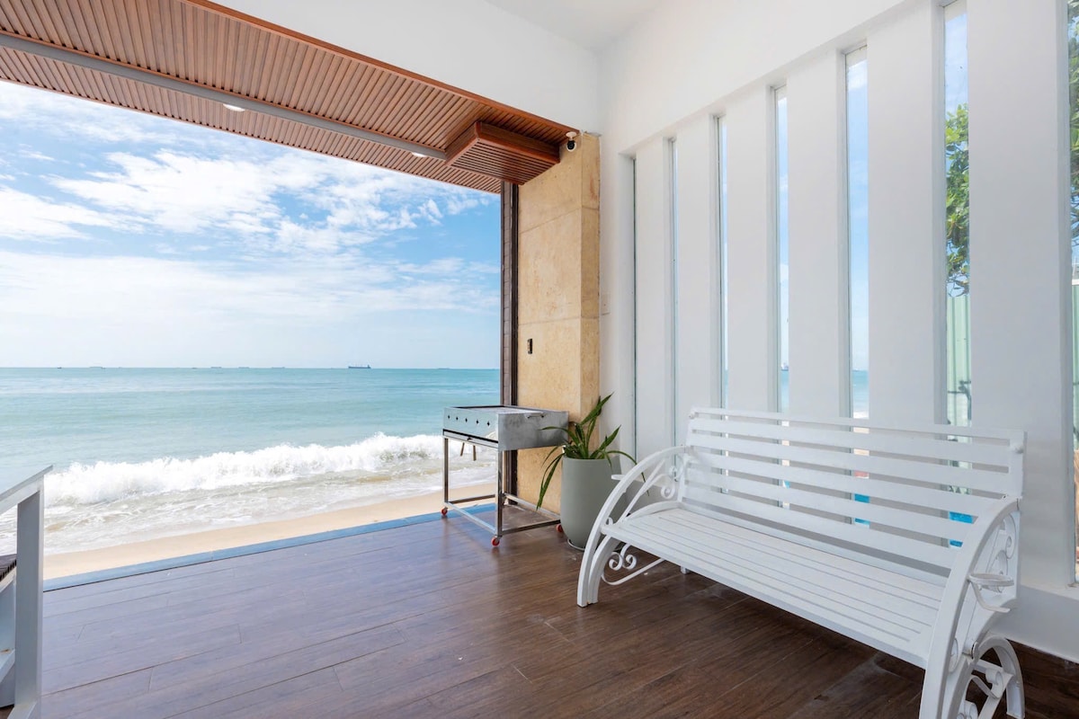 A bright and airy space showcases a white bench positioned near an open glass door, offering views of the ocean waves. Light wooden flooring complements the light-colored walls, and a small grilling station can be seen against the wall, adding functionality to the area.