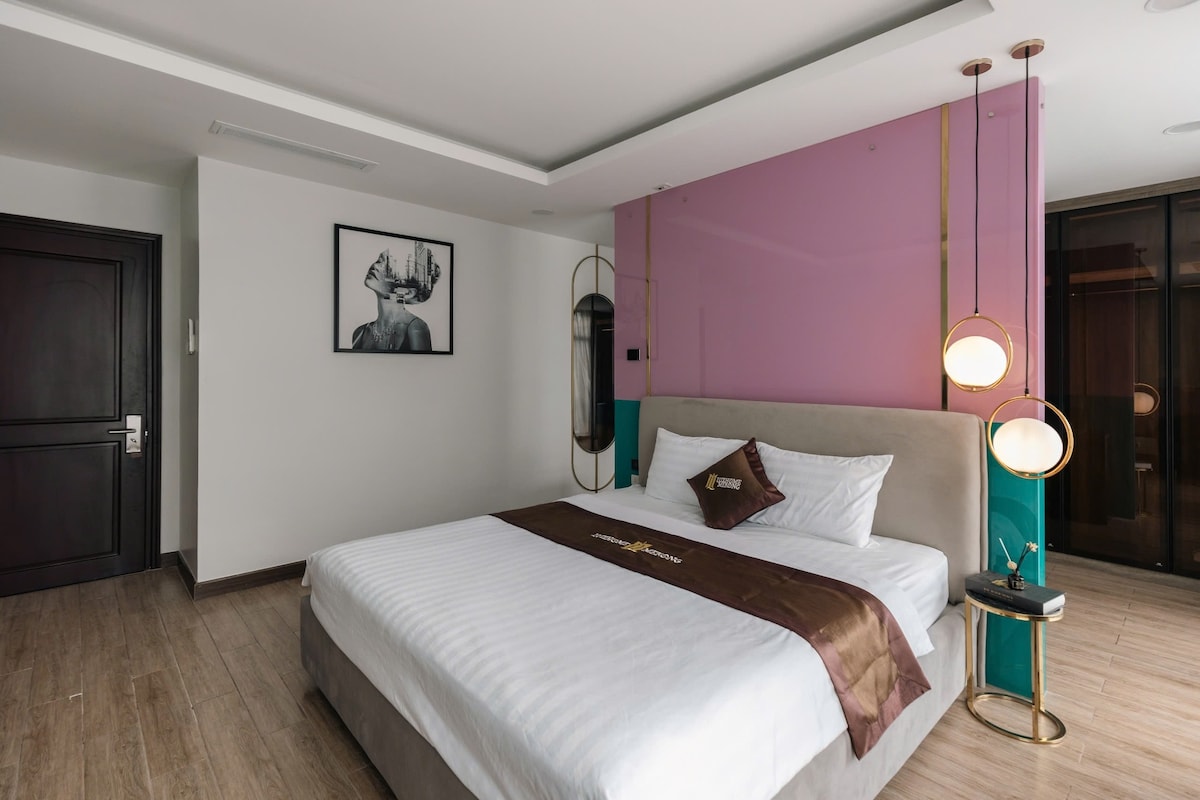 A spacious bedroom is highlighted by a soft bed dressed in crisp white linens. A decorative accent wall in hues of pink and teal adds visual interest. A modern piece of artwork is displayed on the wall, and elegant light fixtures provide a warm glow.