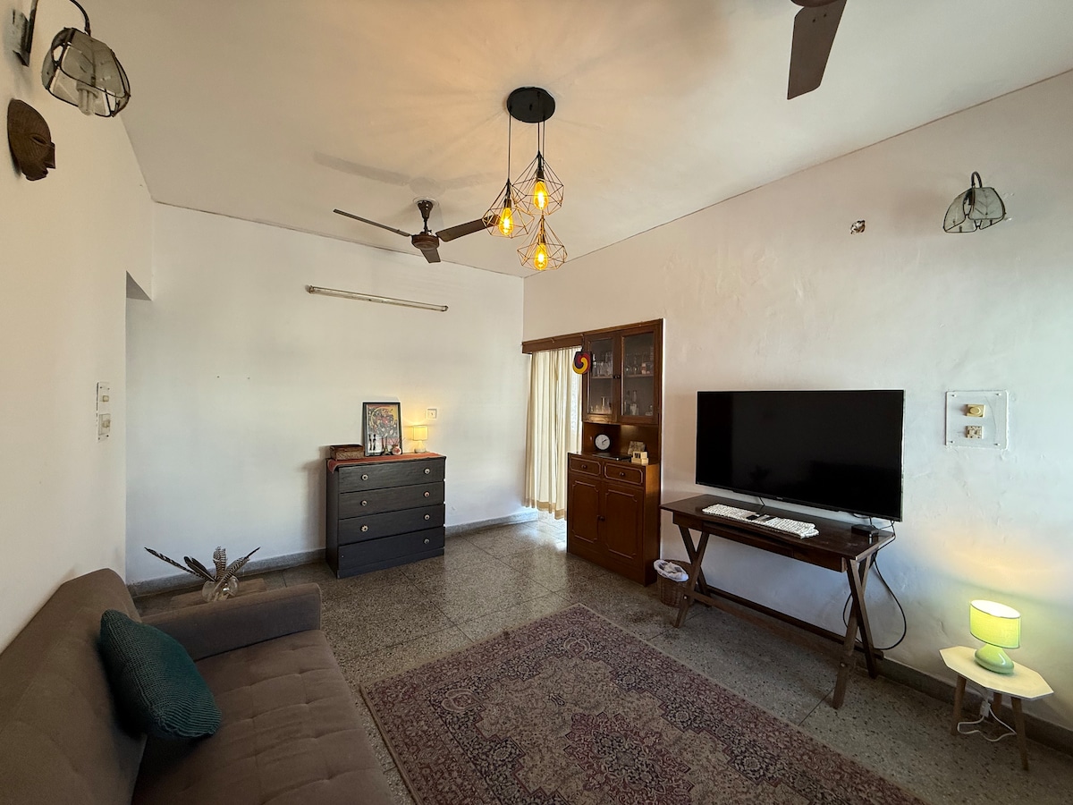 A spacious living room features a comfortable sofa and a wooden cabinet. A flat-screen television is mounted on the wall, and a decorative rug adds warmth to the room. Large windows allow natural light to fill the space, accompanied by various light fixtures.