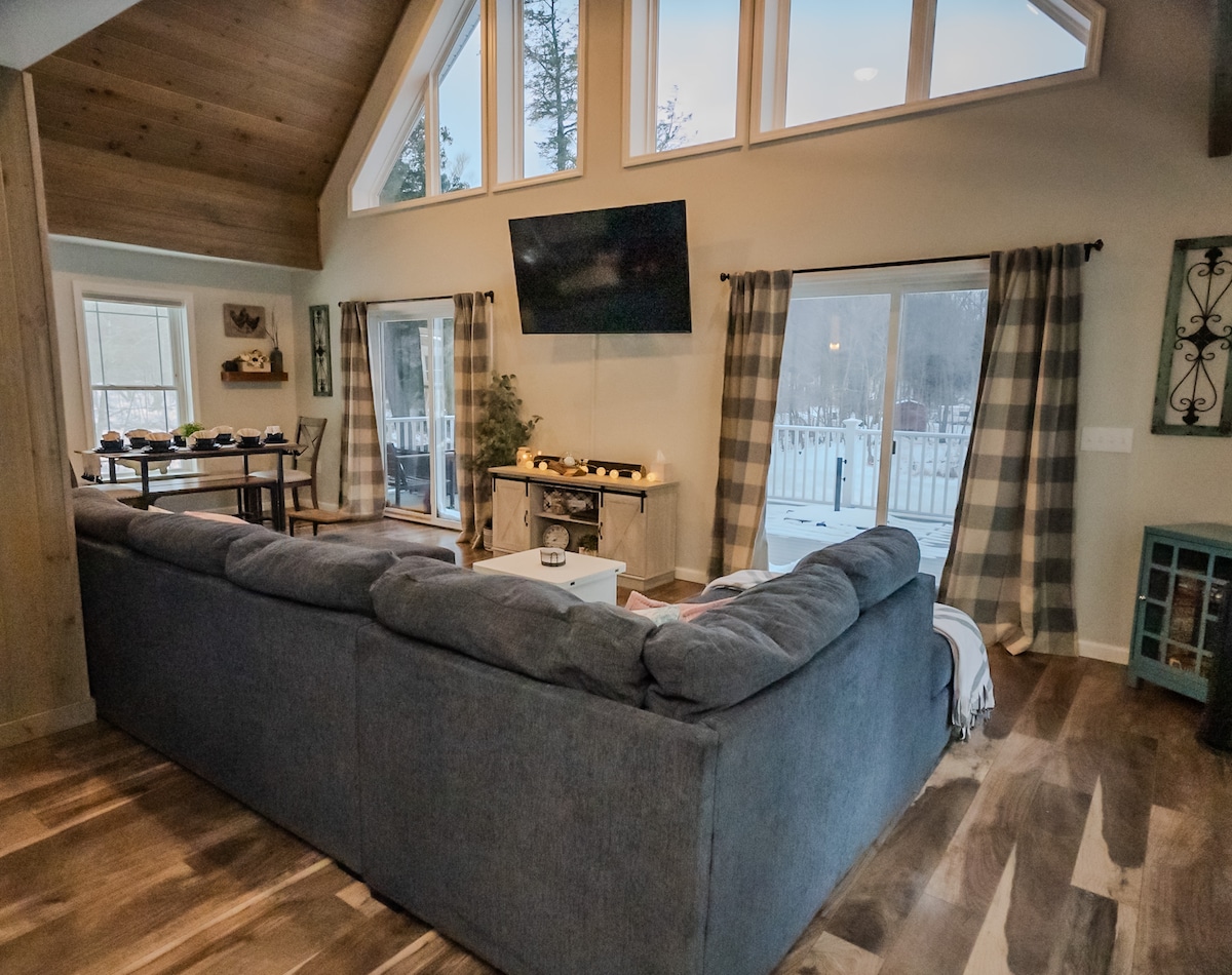 A spacious living area features a large sectional sofa, positioned to face a mounted television. Natural light enters through multiple windows adorned with patterned curtains. A coffee table and side tables complement the cozy setting, with a subtle decor arrangement visible in the background.