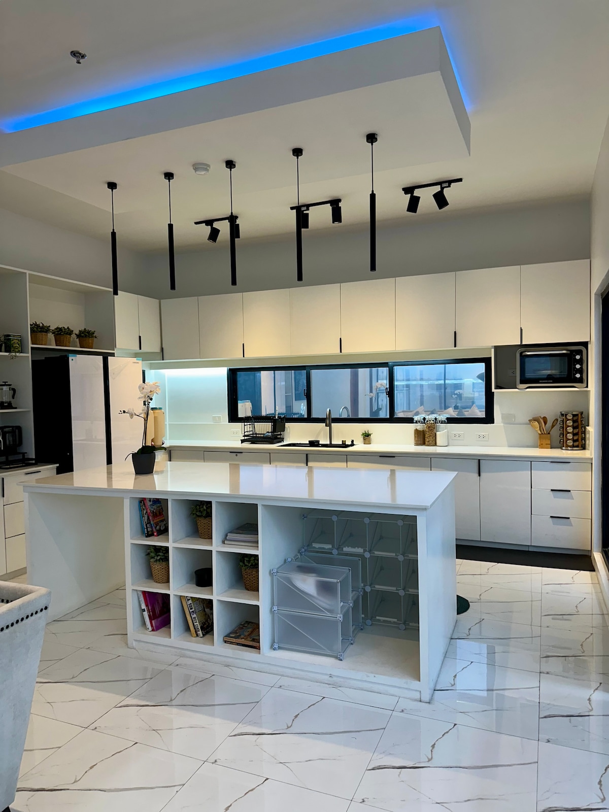 A modern kitchen features sleek cabinetry and a central island with open shelving displaying books and decorative items. Ample counter space is available, with stainless steel appliances and large windows providing natural light and city views.