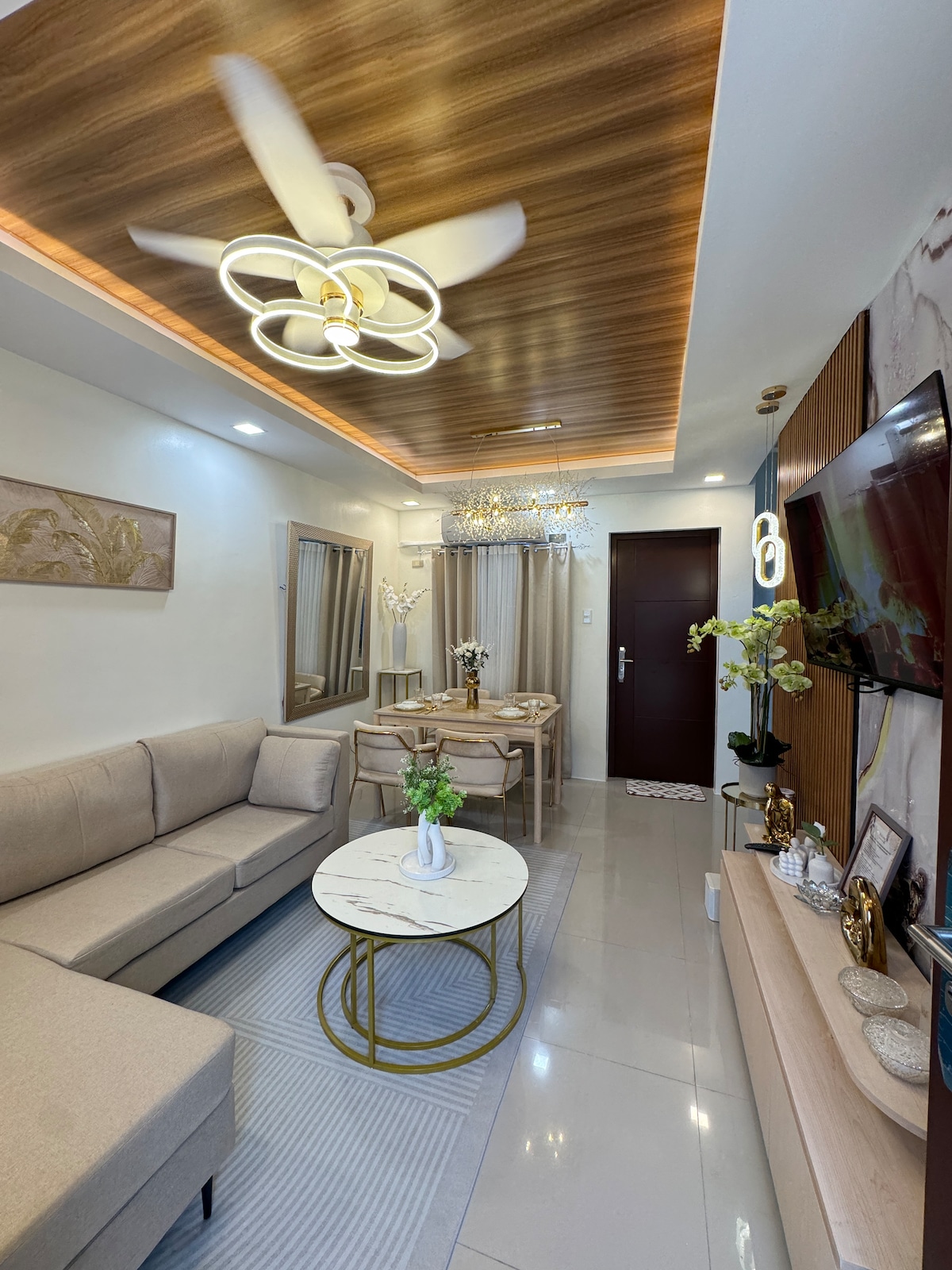 A modern living area features a light-colored sectional sofa and a round marble coffee table. A dining table is set for four, and a ceiling fan and chandelier provide overhead lighting. Reflective surfaces and light-colored walls enhance the room's spacious feel.